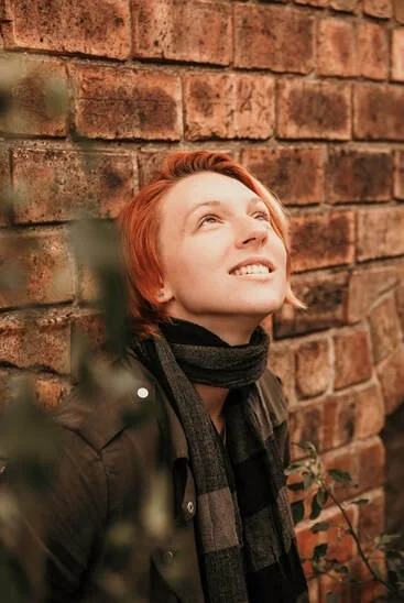Young woman with short red hair smiling and looking upward, standing against a brick wall with green foliage in the foreground.