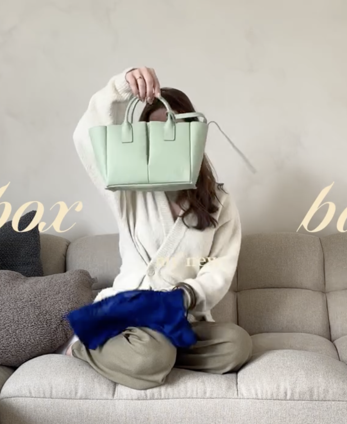 Vrouw zittend op een beige bank, met een groene tas over haar hoofd, een stuk geblokkeerd door haar hand, en een blauw item in haar schoot.