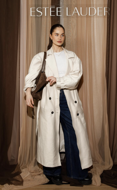 Een vrouw poseert voor een backdrop met de tekst 'Estée Lauder'. Ze draagt een lange, crèmekleurige trenchcoat, blauwe jeans en zwarte schoenen, en houdt een bruine tas vast.