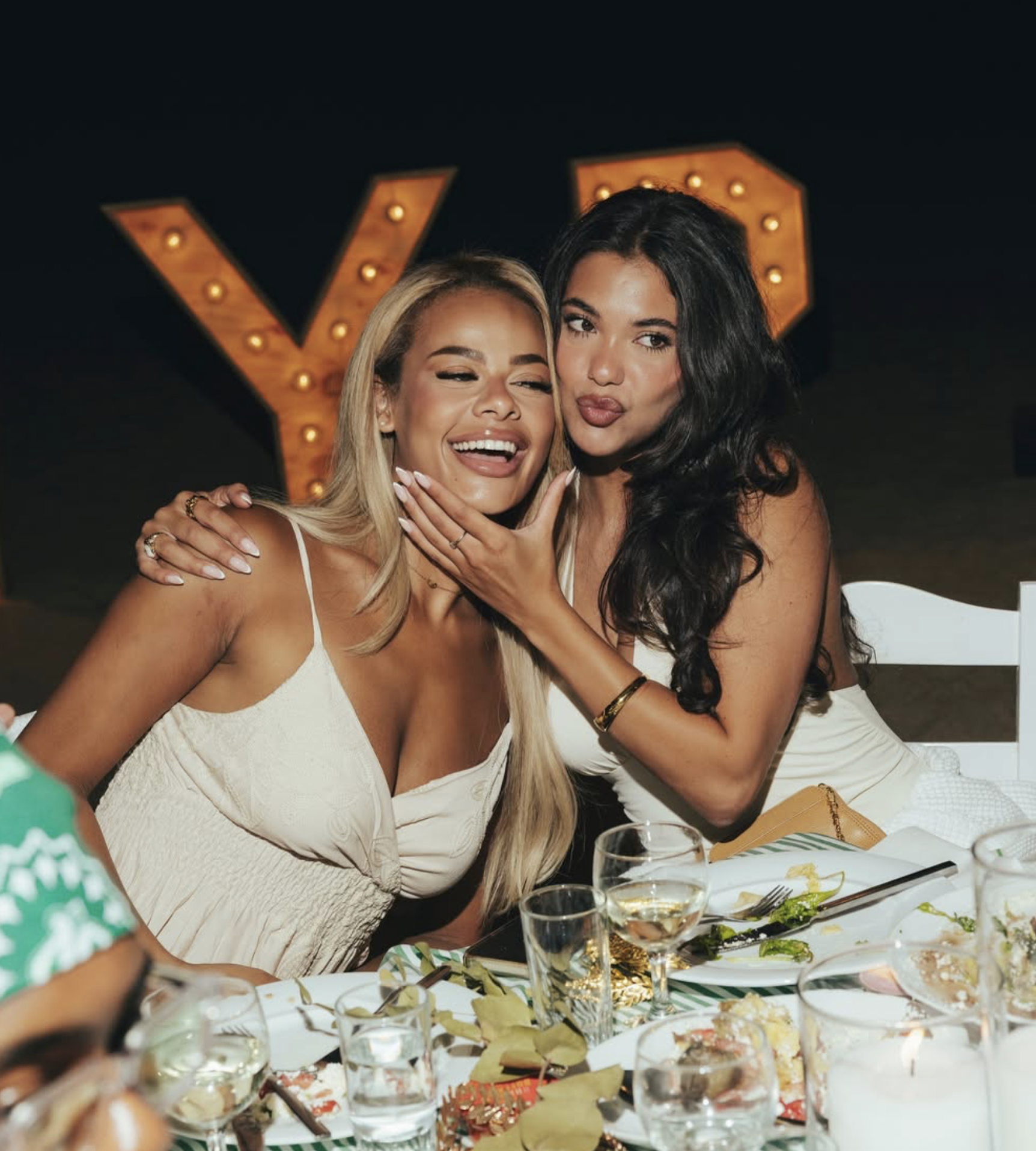Twee jonge vrouwen die samen lachen en poseren op een feestje, met een feestdecoratie en eten op tafel.