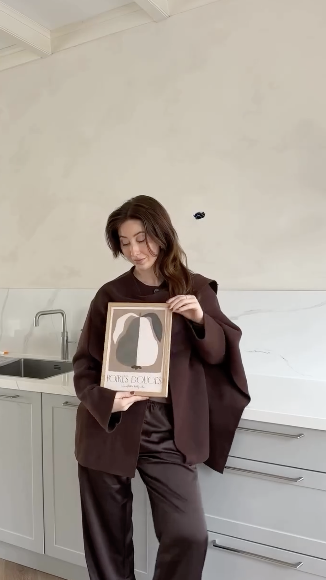 Vrouw in een keuken houdt een poster vast met een illustratie van een perzik en de tekst 'POIRES DOUCES'.