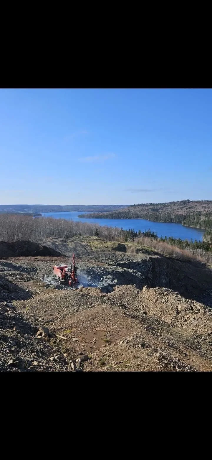 Construction site on a hillside with a red excavator working, overlooking a large river and a forested landscape under a blue sky.