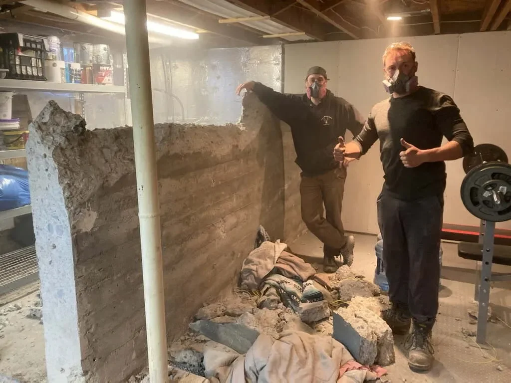 Two men wearing masks in a basement or garage, standing next to a large broken concrete wall with visible brickwork behind, giving a thumbs up and pointing at the wall. There are construction supplies and a weightlifting bench nearby.