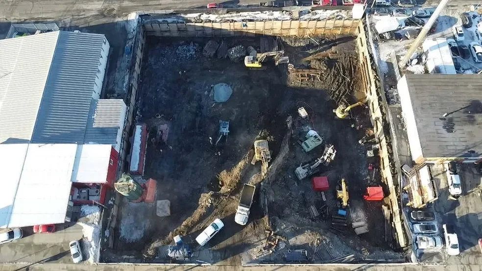 A construction site viewed from above with excavation equipment and several vehicles inside the area, surrounded by buildings and parked cars.