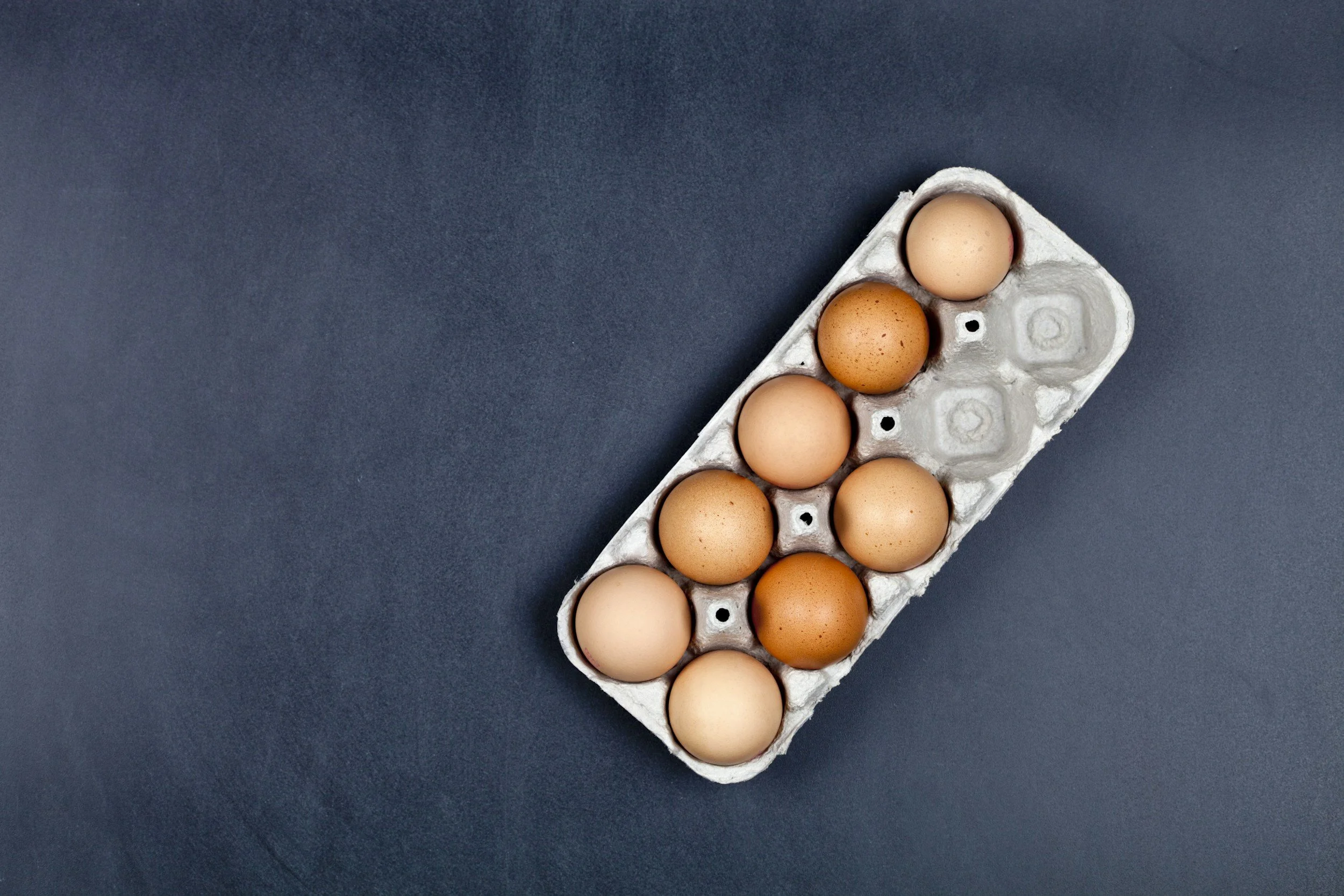 Una bandeja de cartón con varios huevos de diferentes tonalidades de marrón, sobre un fondo gris oscuro.