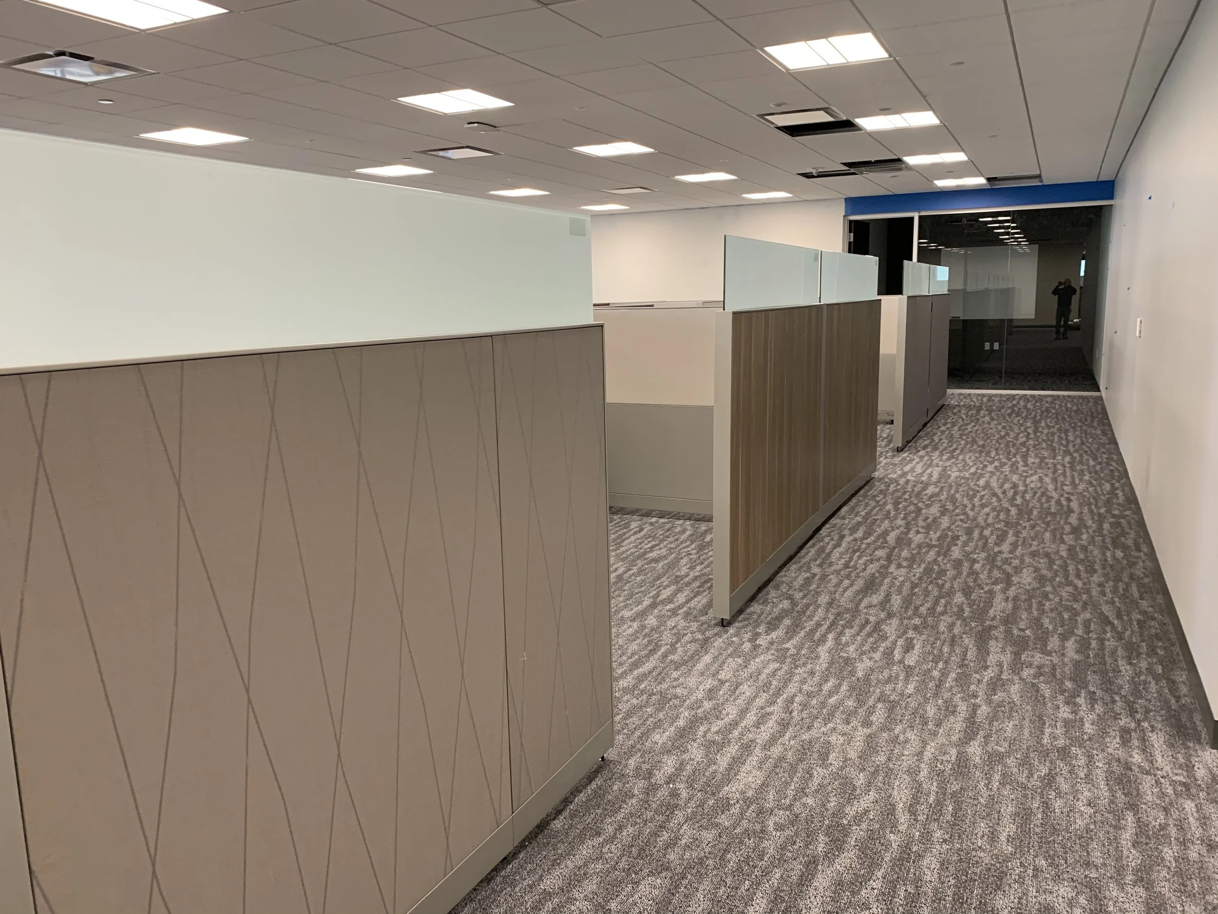 Empty office space with cubicles, white walls, gray patterned carpet, and a ceiling with fluorescent lights. A person is visible in a reflection in the background taking a photo.