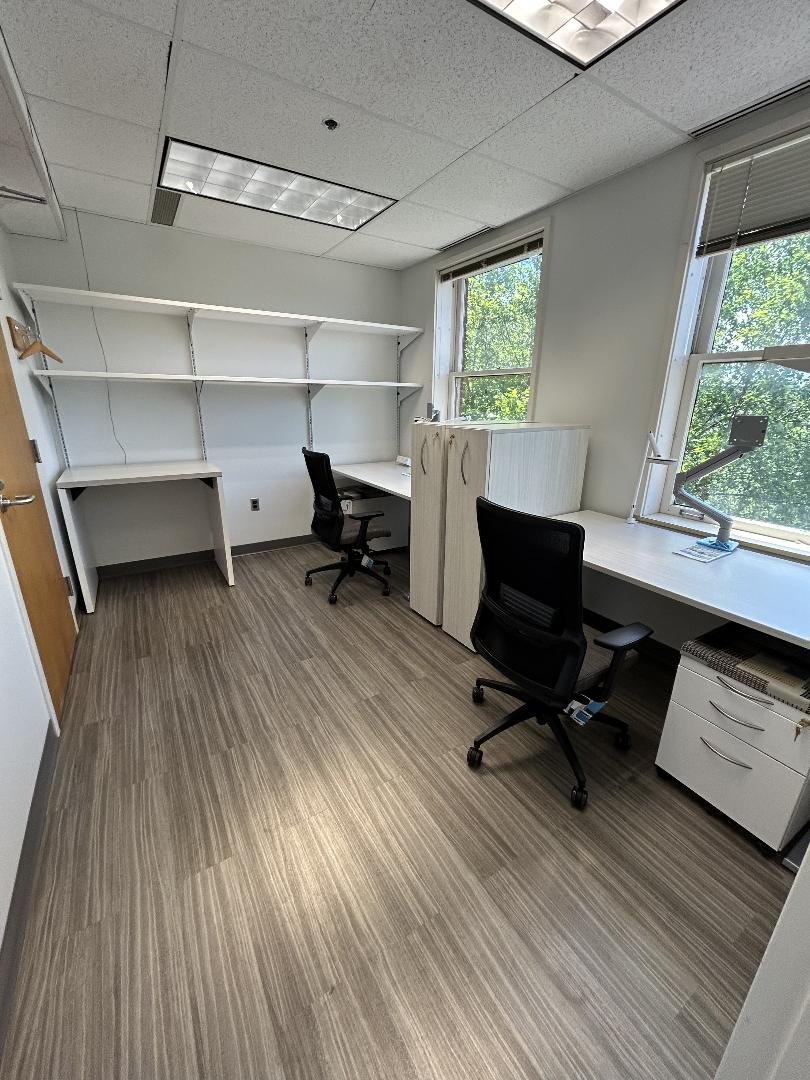 An empty office with white walls, wood flooring, two desks with black office chairs, large windows, and white shelving on the back wall.
