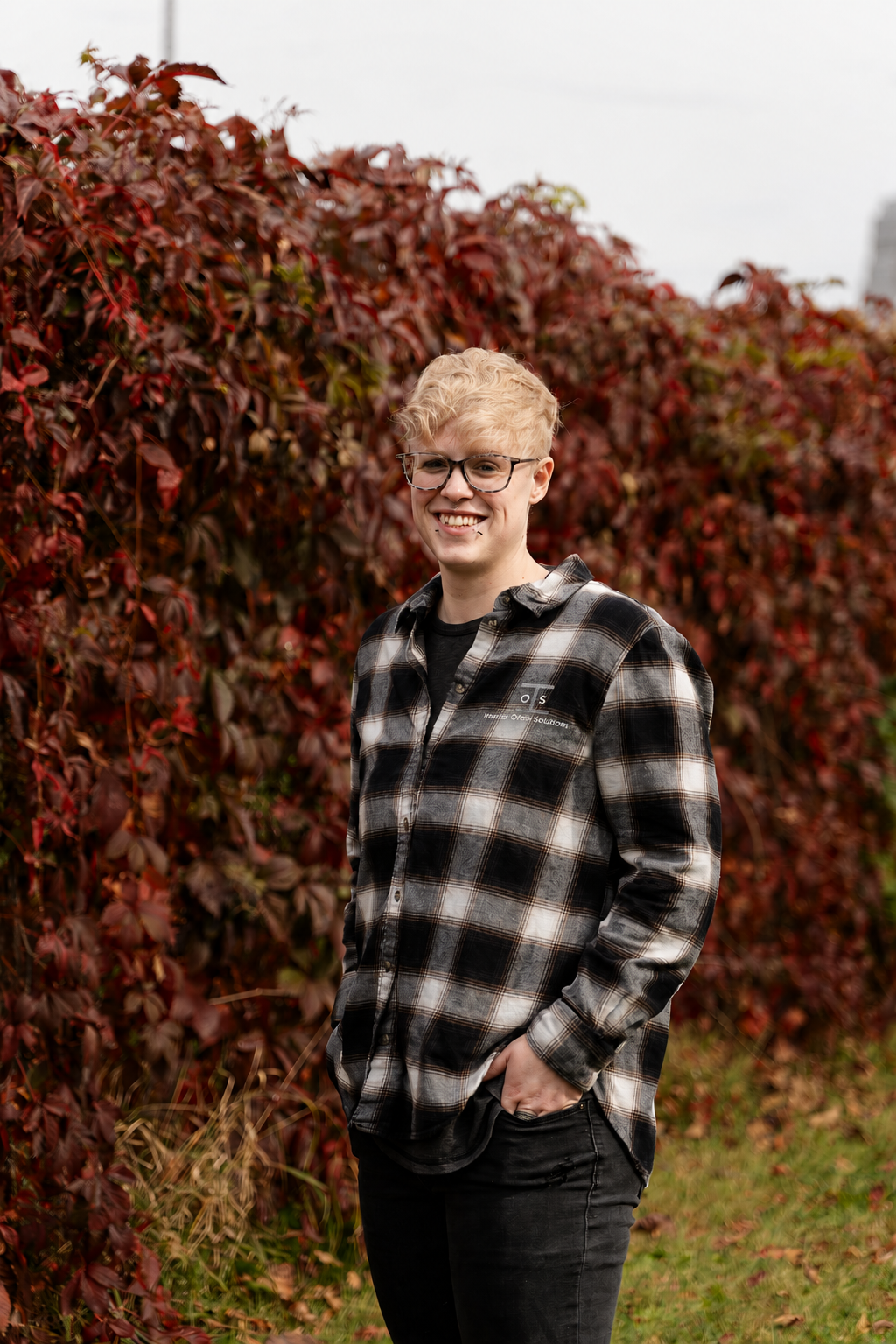 A smiling young person with blonde hair, glasses, and a black and white plaid shirt standing outdoors in front of a red, leafy hedge during daytime.