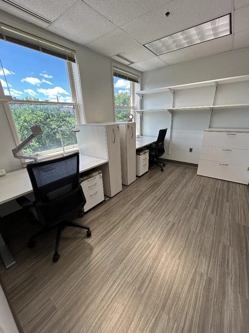 Empty office space with two desks, office chairs, large windows, and built-in shelves.