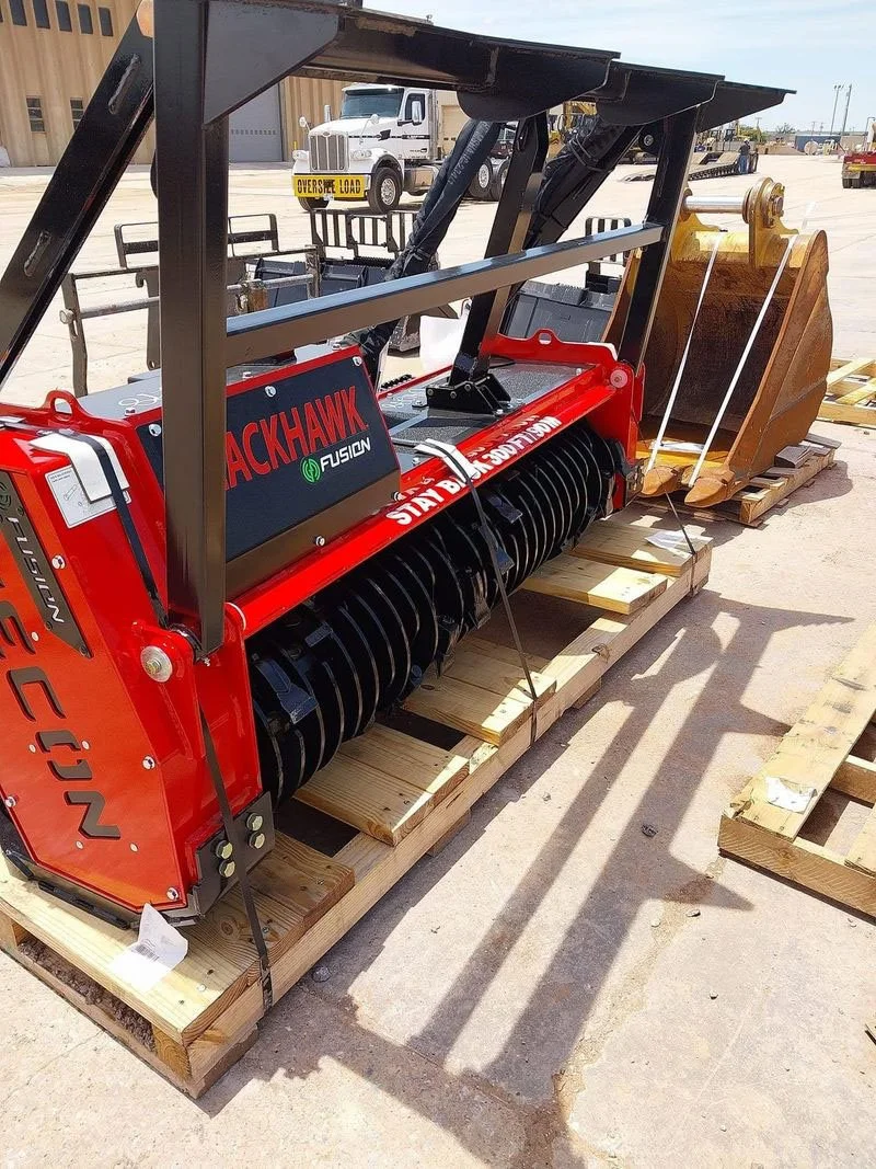 Red and black Bobcat compact track loader attachment on wooden pallets, with a large bucket at the front, placed outdoors in a construction site. There is a tanker truck and construction materials in the background.