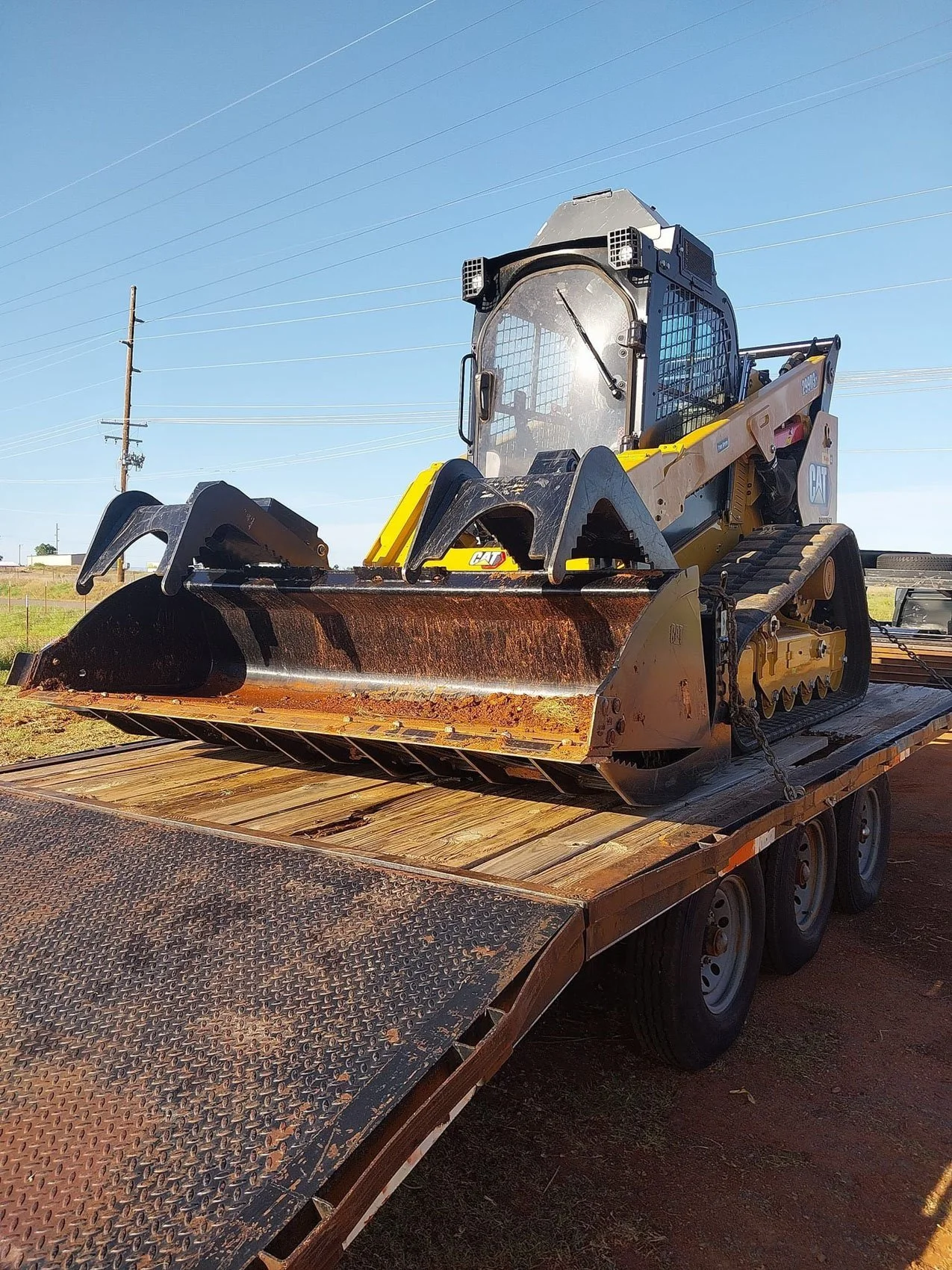 A compact bulldozer on a flatbed trailer with wooden and metal surface, outdoors under a blue sky, power lines in the background.