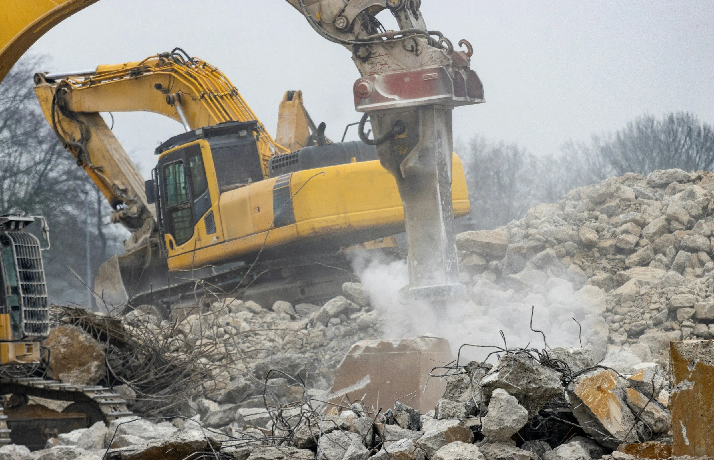 A yellow construction excavator breaking rocks on a construction site with dust and debris in the air.