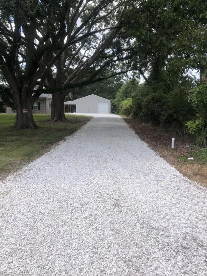 Gravel driveway leading to a white garage surrounded by trees and lawn.