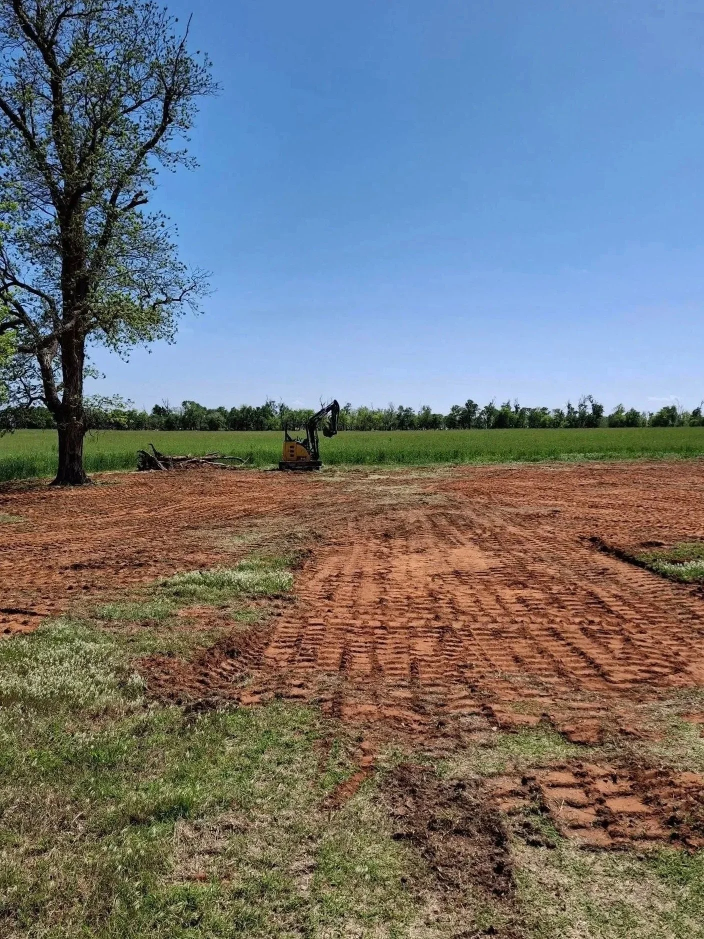 A dirt path with tire tracks runs through a grassy field toward a line of trees at the horizon. To the left, a large tree stands, and a small excavator is parked near the tree on the dirt area. The sky is clear and blue.