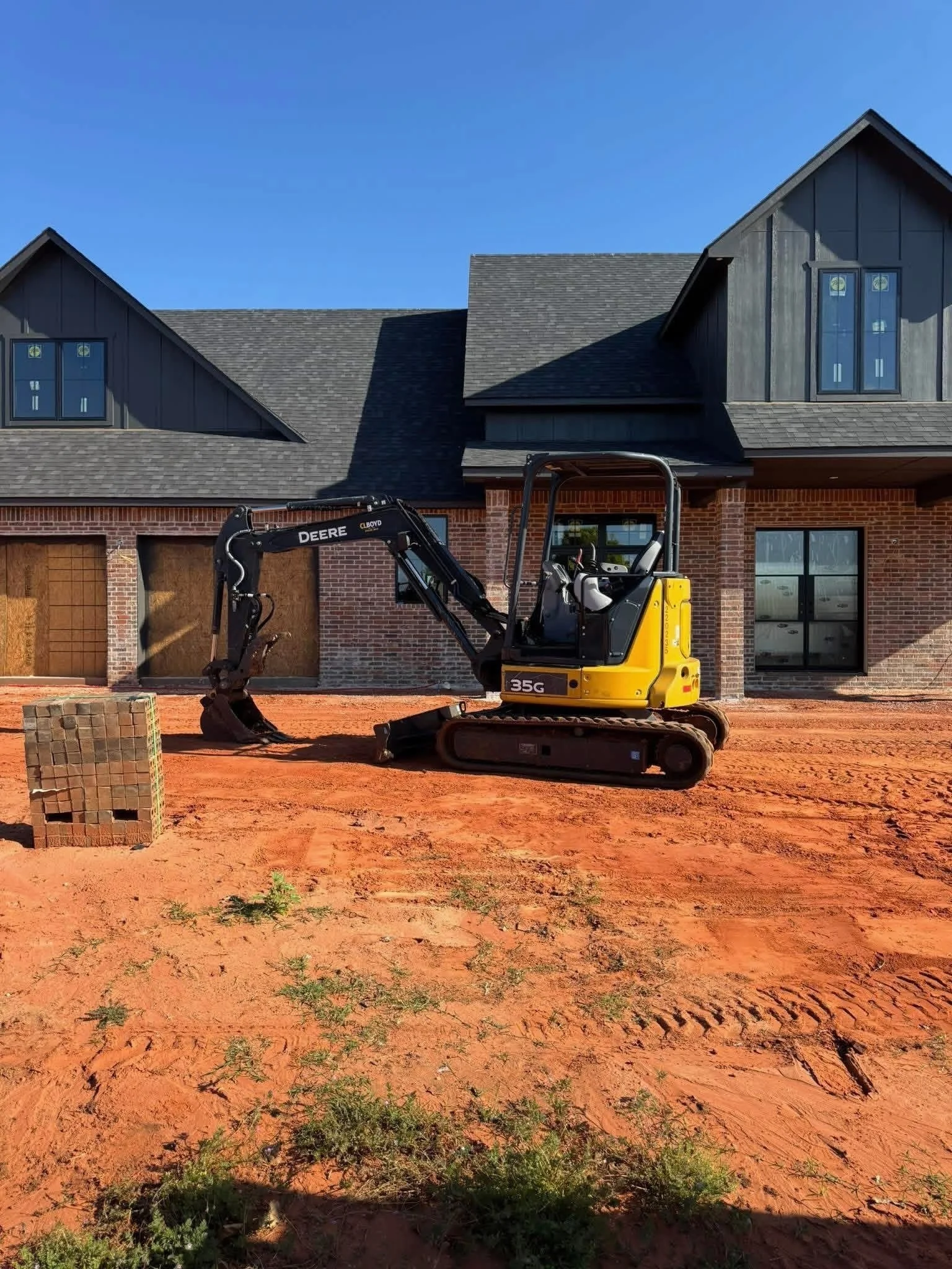 Construction site in front of a house with a black roof, red brick exterior, and large windows, with a small yellow excavator on the dirt ground.