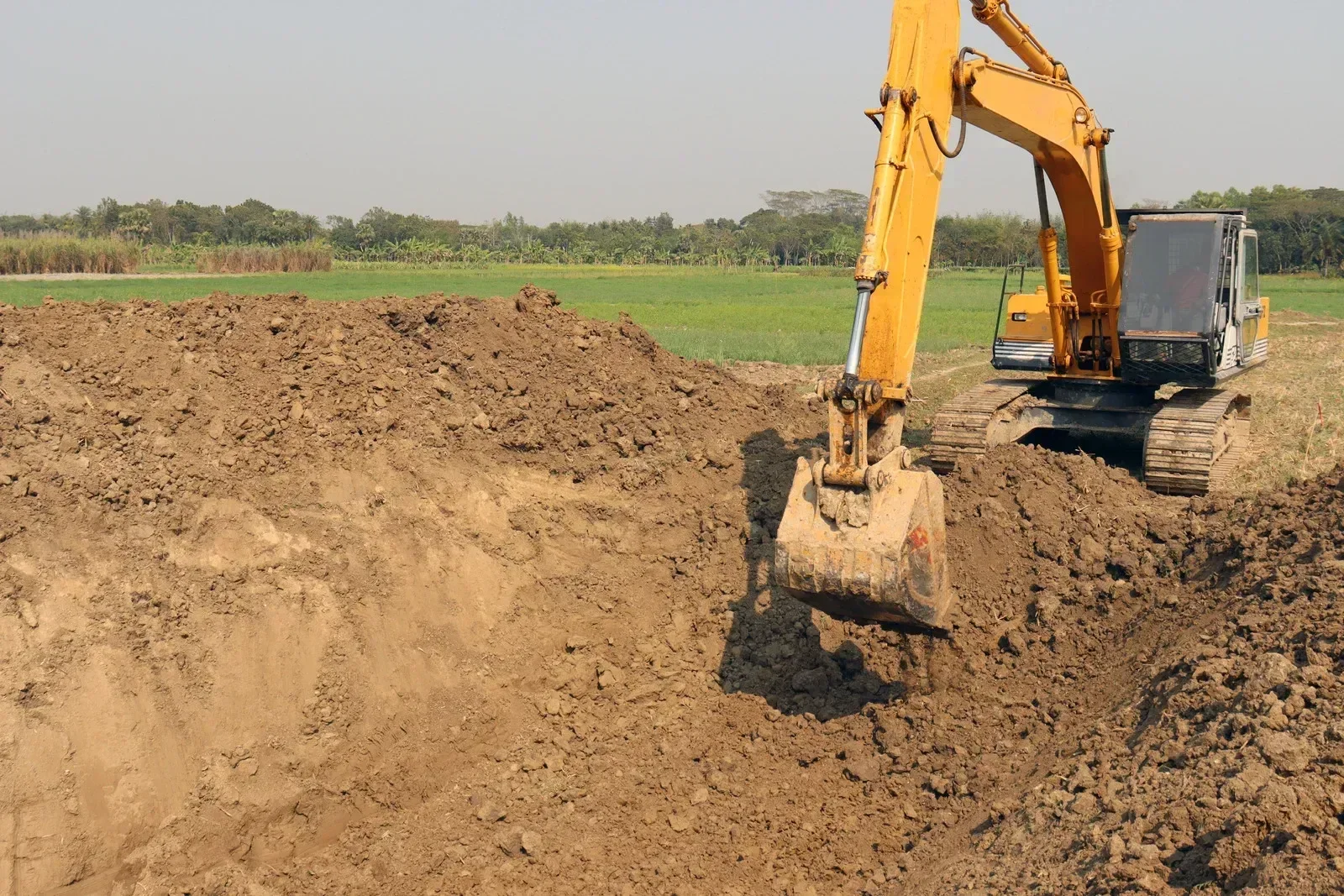 A yellow excavator moving dirt in a field with greenery and trees in the background.
