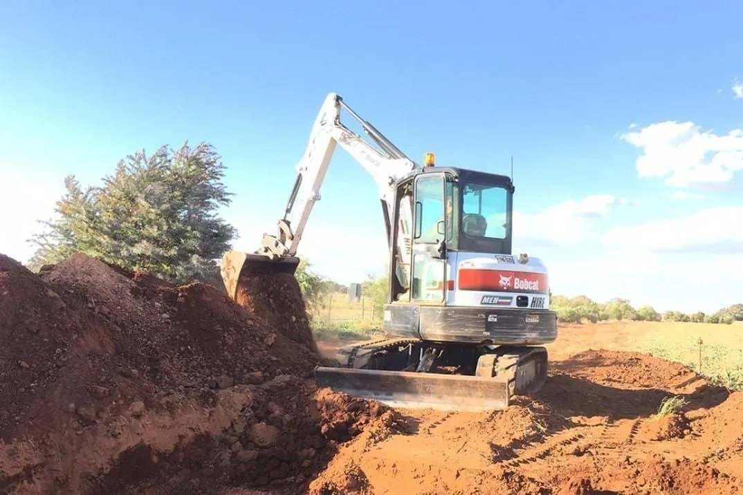 Excavator moving dirt in a construction site with open field and partly cloudy sky in the background.