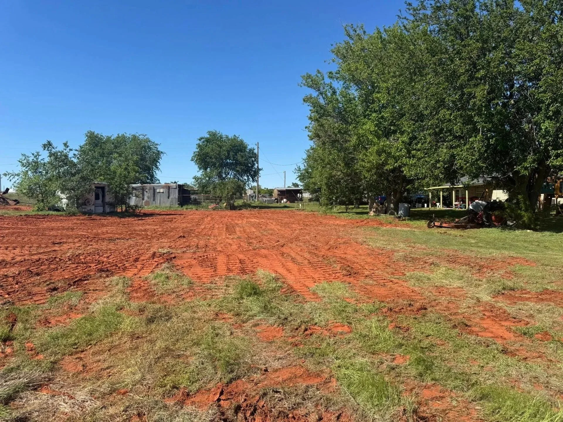 A rural scene with a cleared patch of reddish dirt, bordered by green grass, and several large trees. In the background, there are small buildings or sheds and parked vehicles, with a bright blue sky overhead.