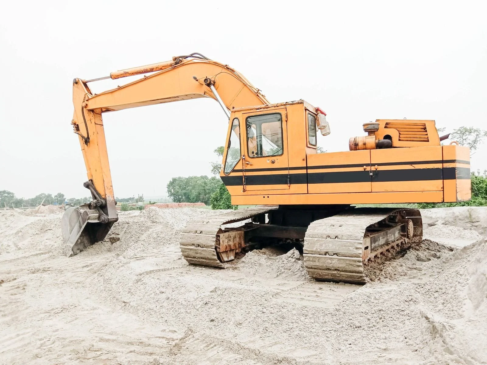 An orange excavator digging into a mound of light-colored sand or dirt at a construction site, with a background of trees and a cloudy sky.