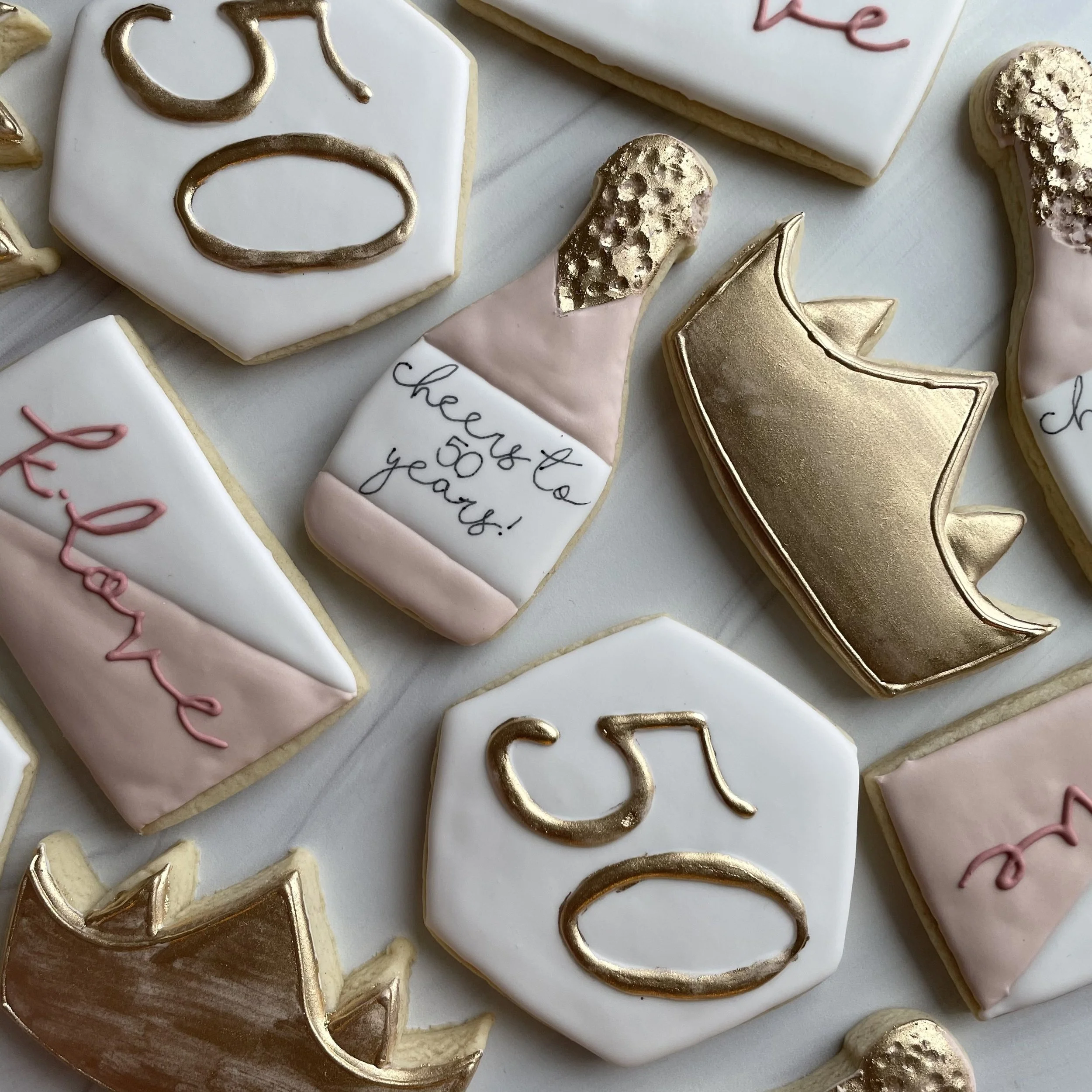 Close-up of decorated cookies with white, pink, and gold icing, some with message writing and others in shapes like stars and moons.