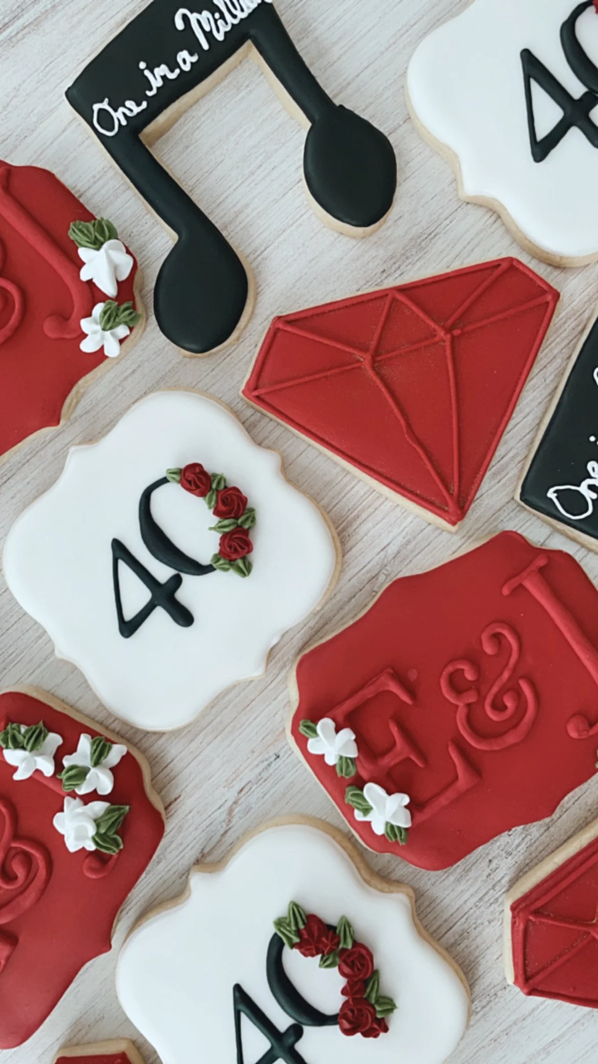 Decorated cookies in various shapes, including a music note, a diamond, and speech bubbles, with red, black, and white icing, some adorned with small flowers and lettering.