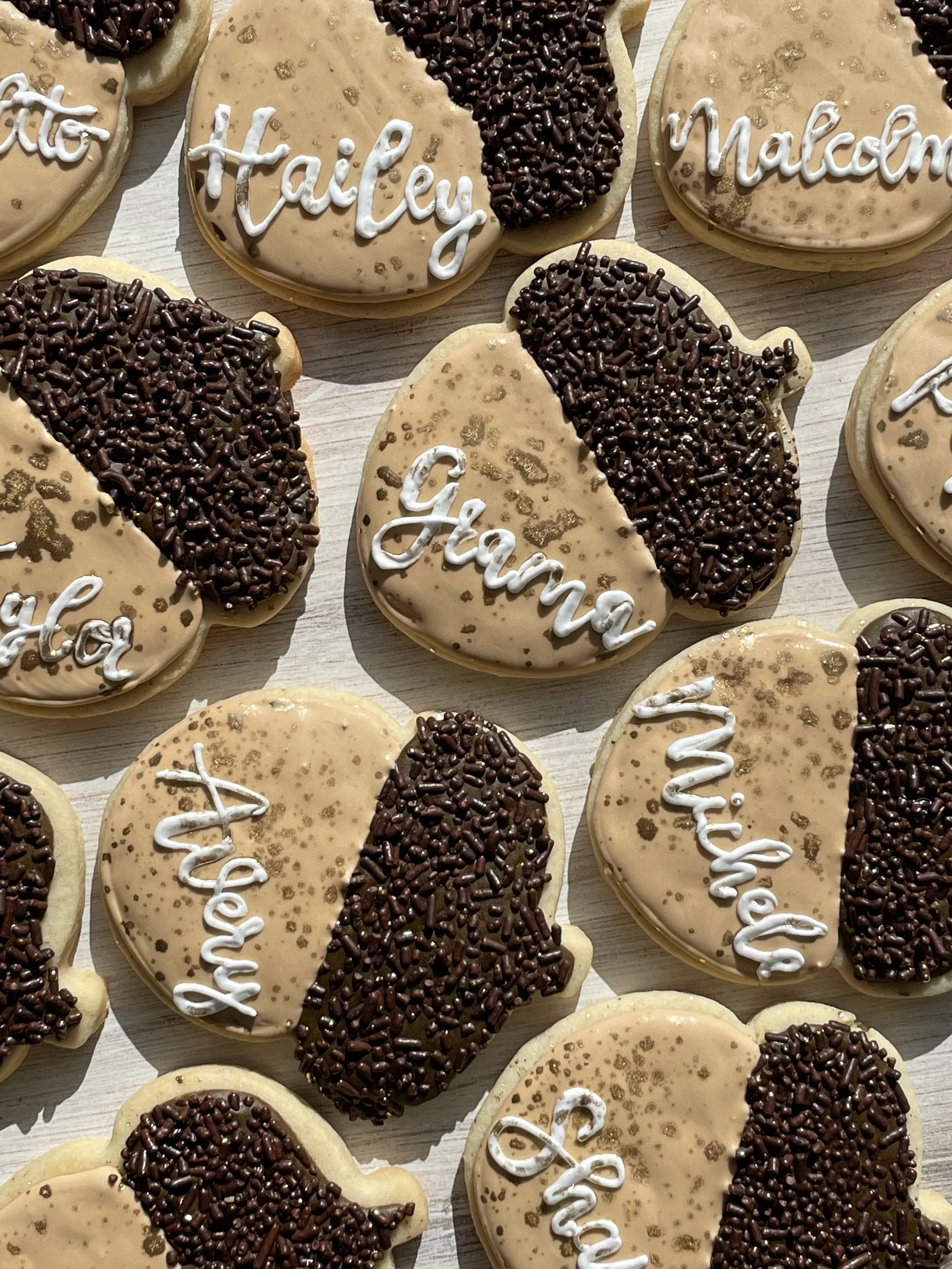 Heart-shaped cookies decorated with beige icing, dark chocolate sprinkles on one side, and white icing writing, with words 'Malcolm', 'Hailey', and 'Maddie' visible.