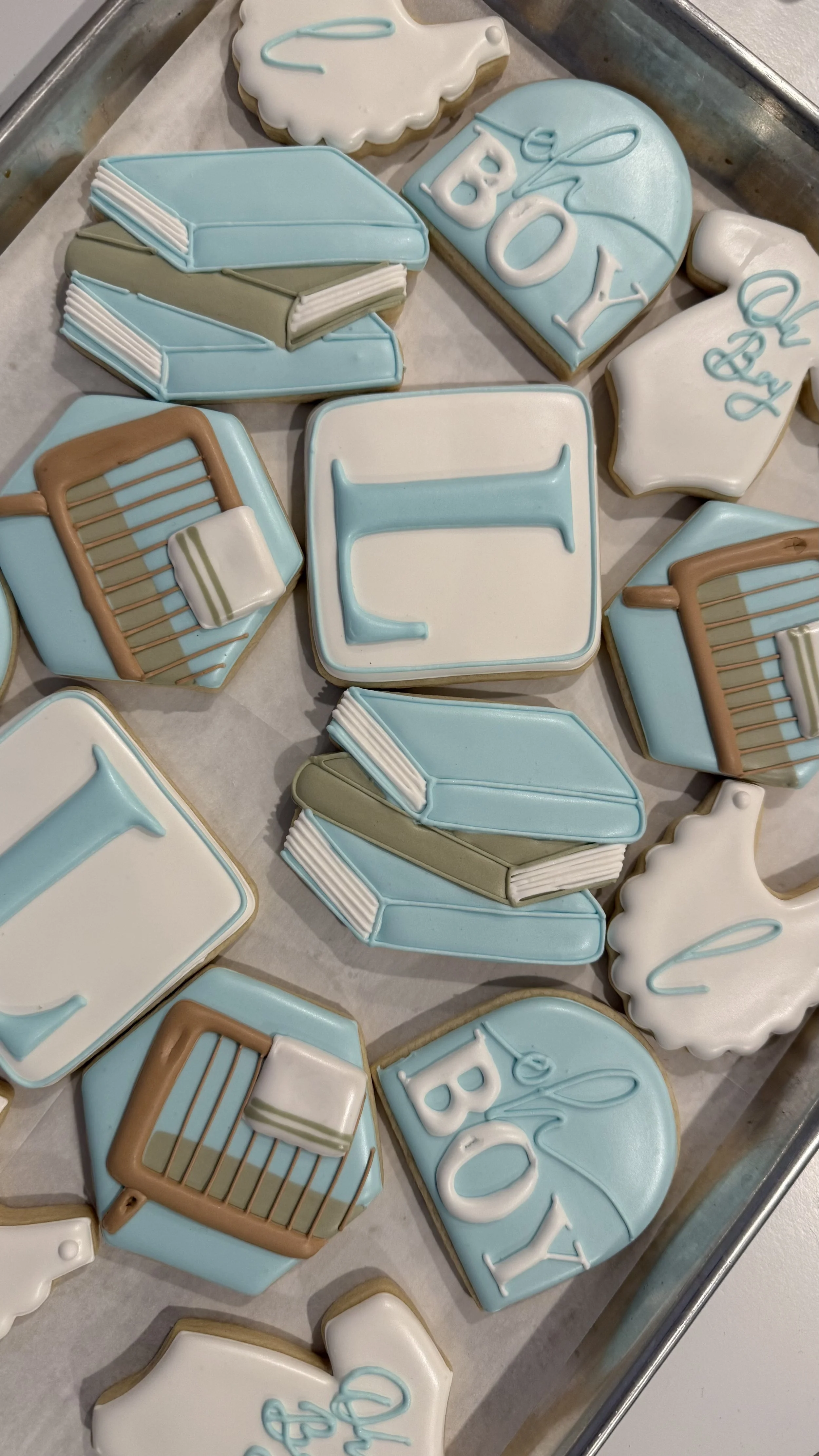Decorated cookies for a baby shower, including diapers, books, a bottle, a crib, and the words "Oh Boy" in light blue and white icing.