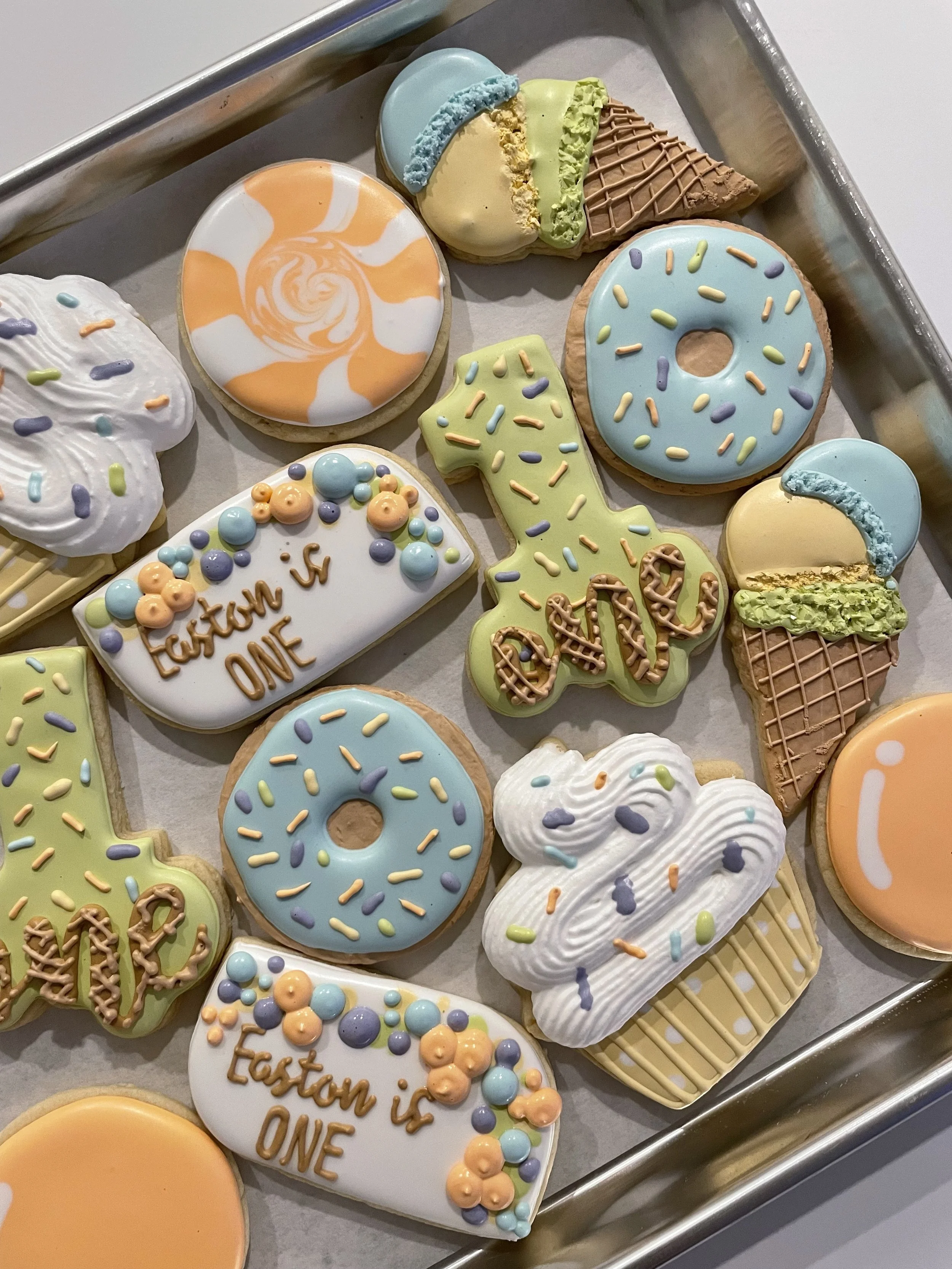 A variety of decorated cookies on a metal tray, including donuts, a lizard-shaped cookie, a triangle-shaped cookie, and a rectangular cookie with colorful sprinkles and the words "Easter is ONE."
