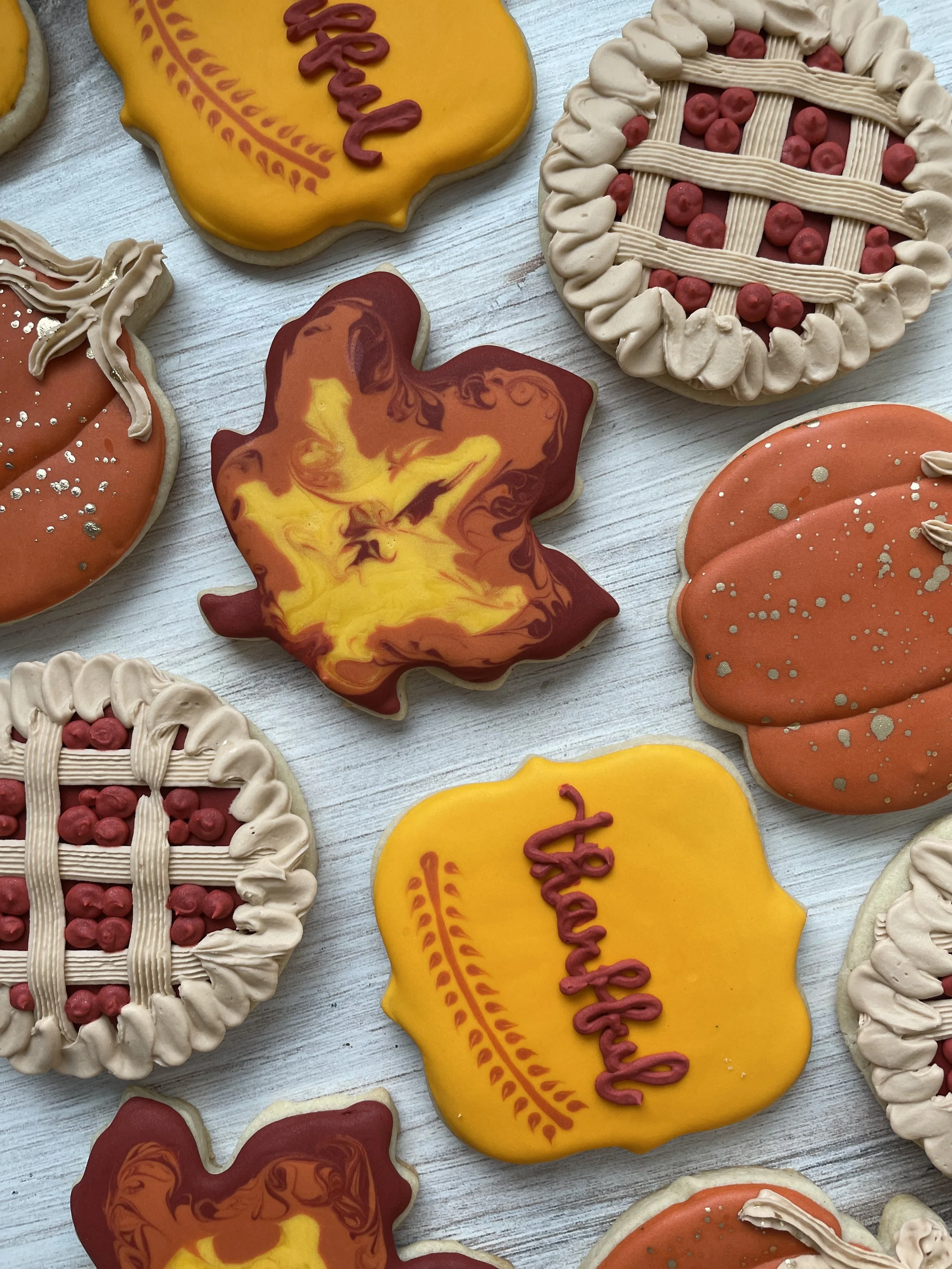 Assorted decorated cookies with Thanksgiving themes, including pie and harvest motifs, on a white wooden surface.