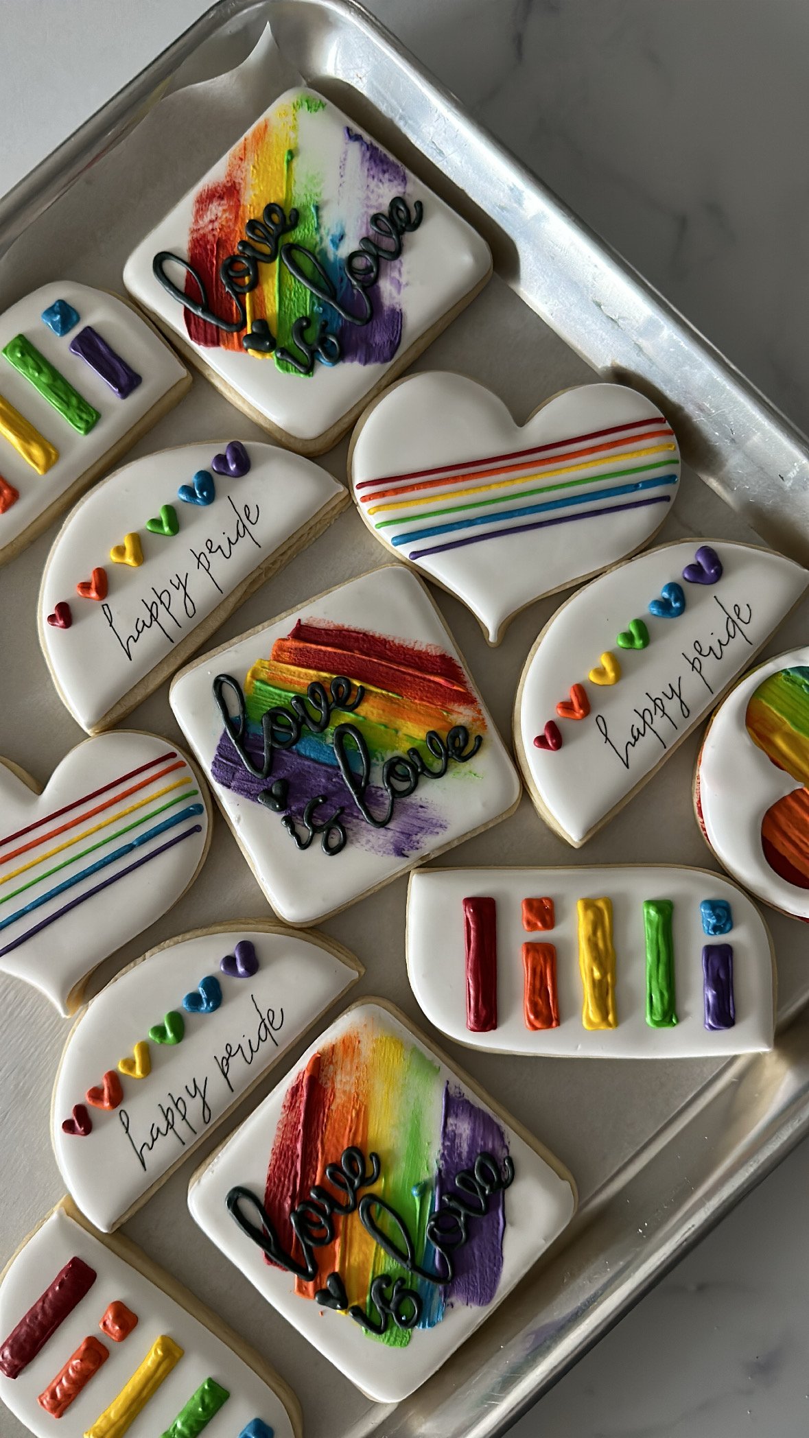 Decorated sugar cookies with rainbow motifs, rainbow-colored lines, and handwritten messages like "happy pride" with colorful hearts, arranged on a silver tray.