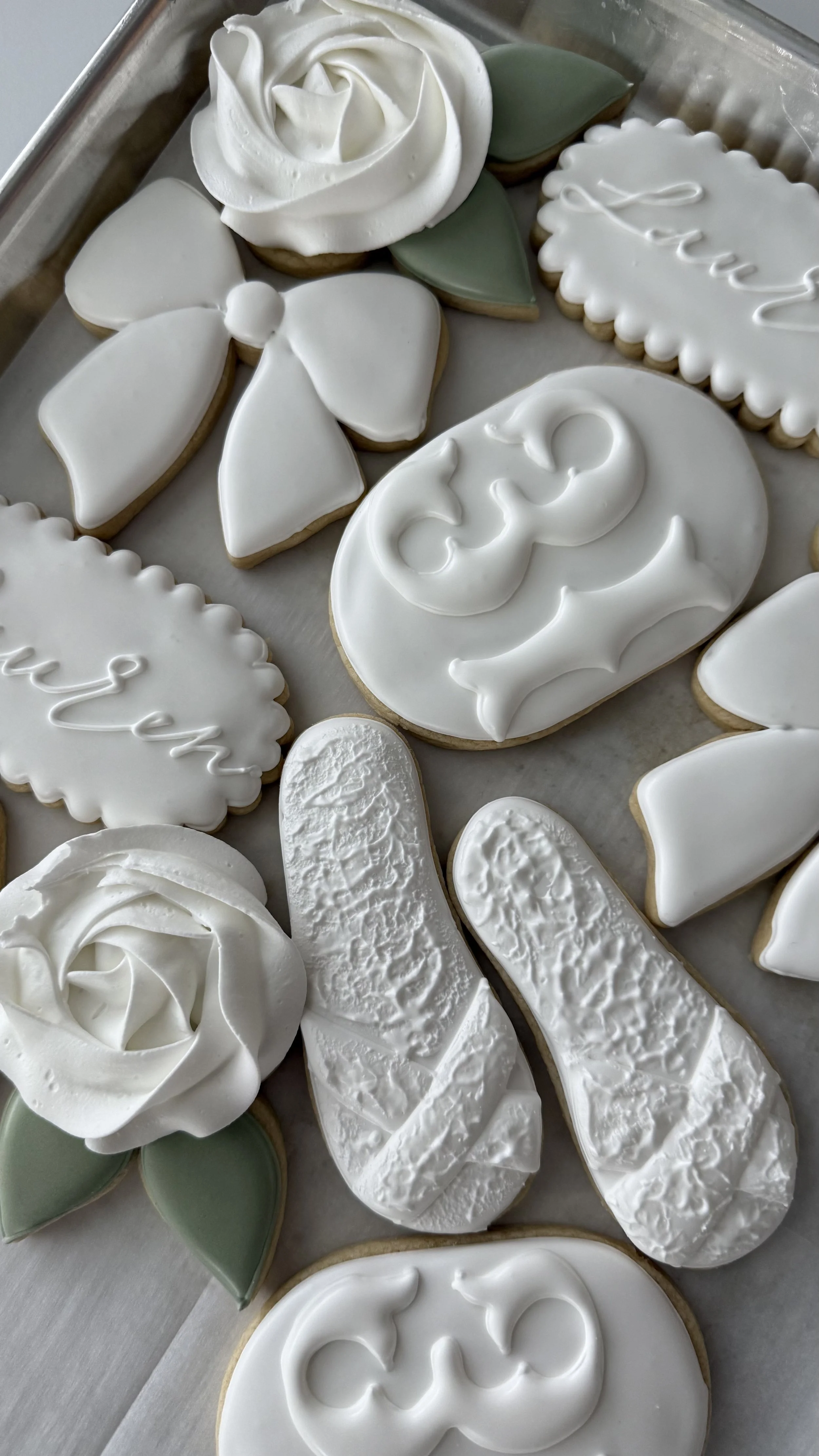 Assorted decorated sugar cookies with white icing, including floral, butterfly, and text designs, on a baking sheet.