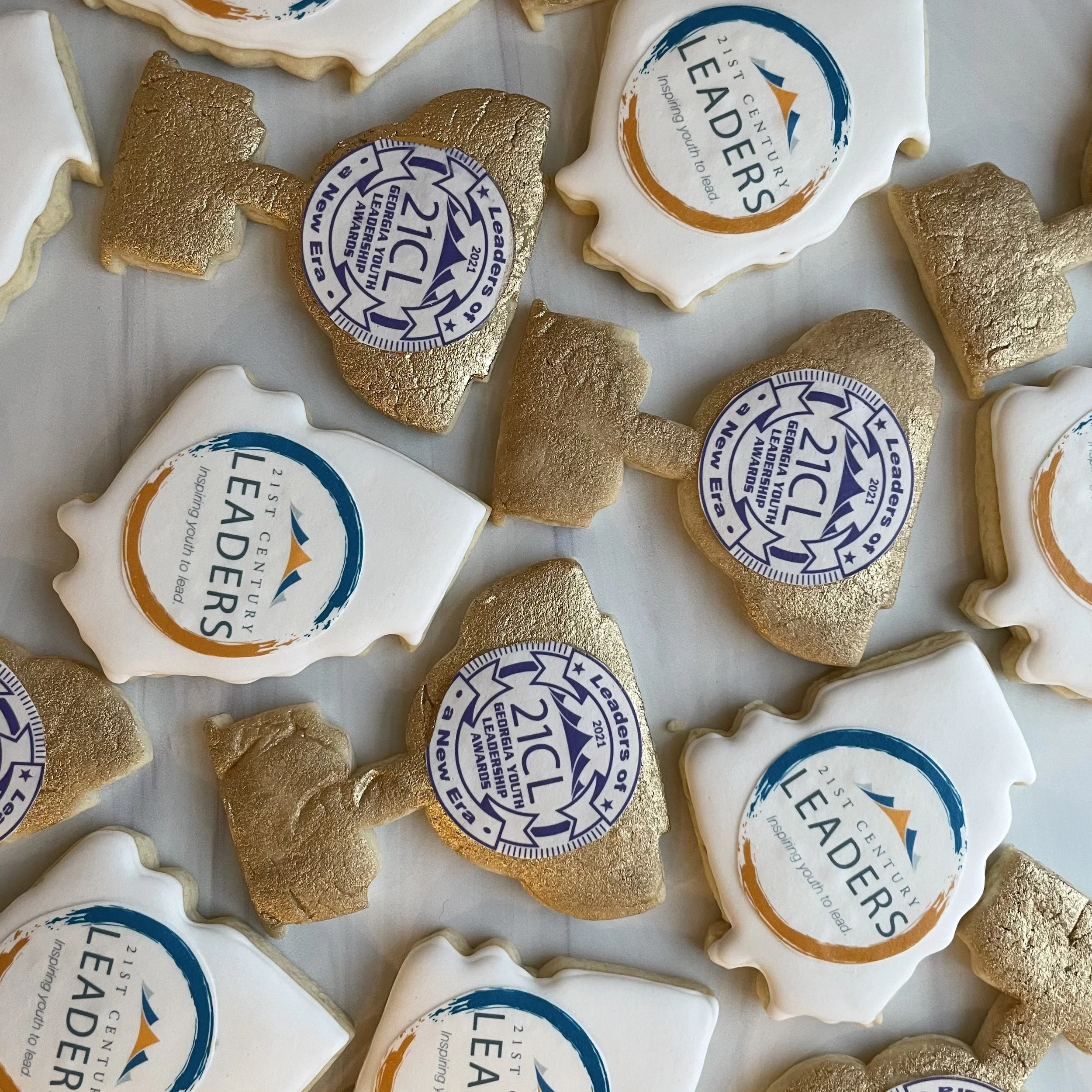 Close-up image of decorated cookies, some with white icing and some with golden edible foil, with circular logo toppers for Georgia Leadership of 211 CICC 2021 and Leaders of Tomorrow.