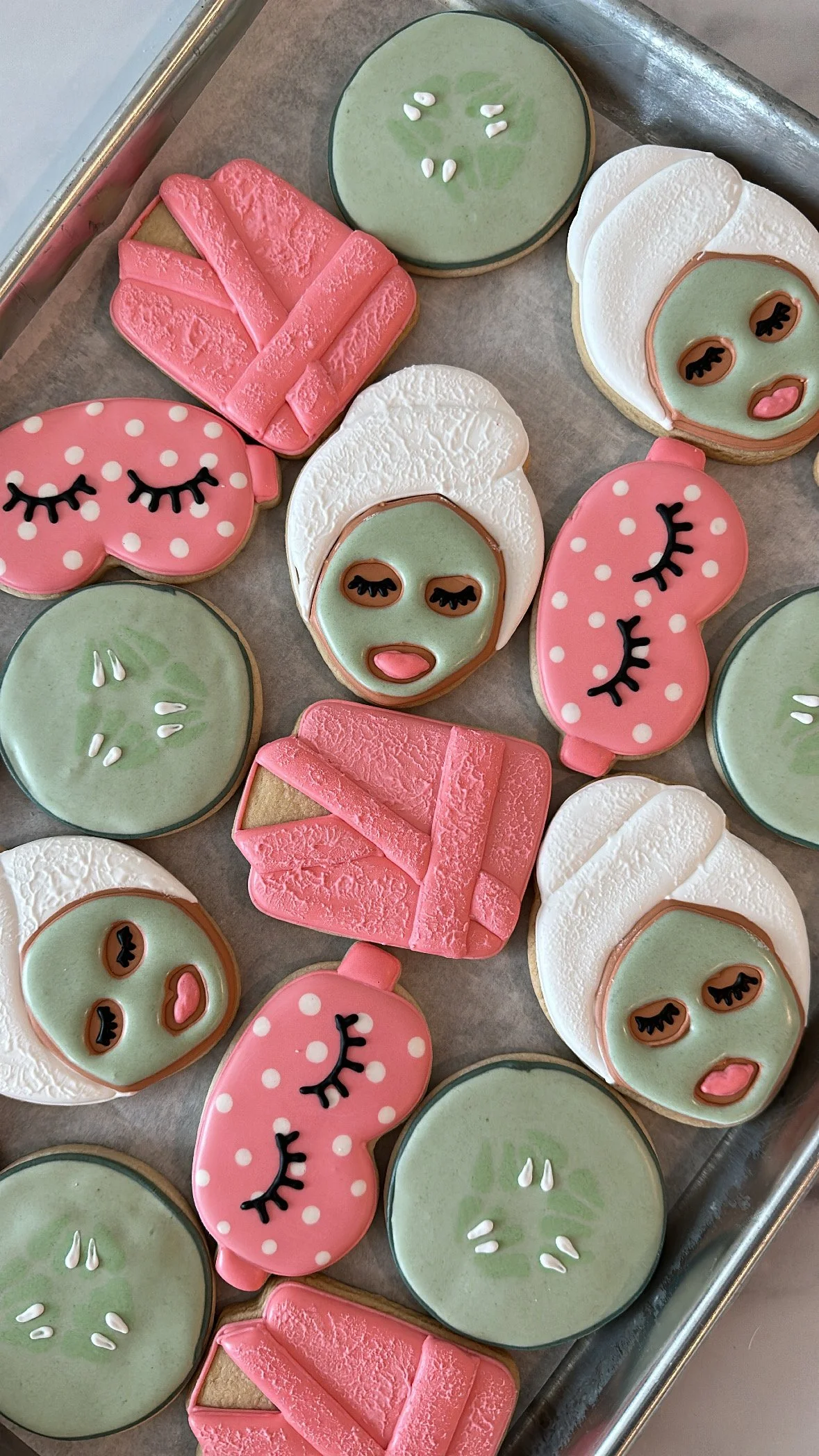 Array of decorated cookies with spa and relaxation themes, including face masks, eyes closed with eyelashes, and pink polka dot cups.