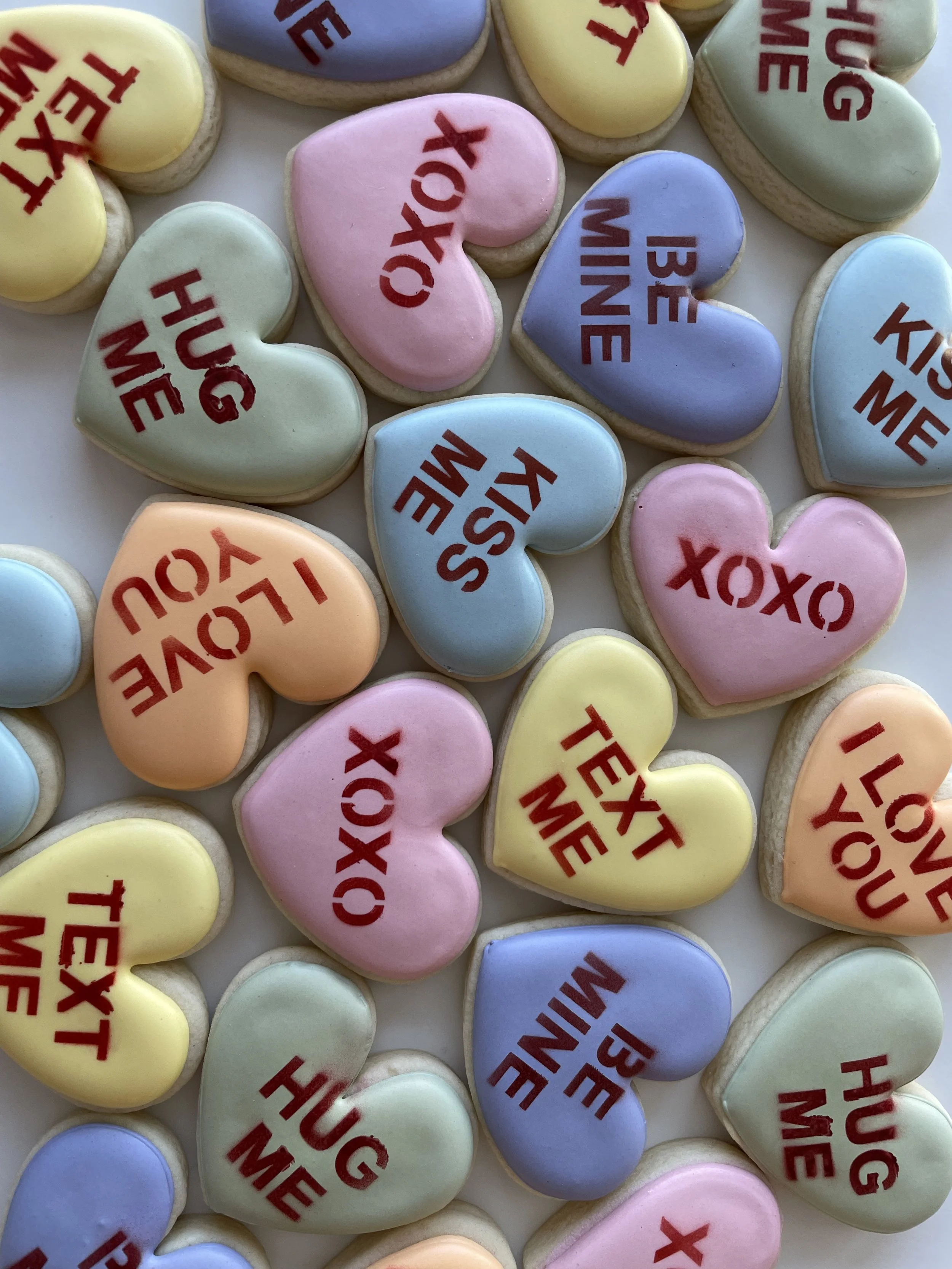 Assorted heart-shaped cookies with pastel icing in pink, yellow, blue, green, and purple. Some cookies have the phrase "HUGS YOU" or "XOX" written in red text on top.