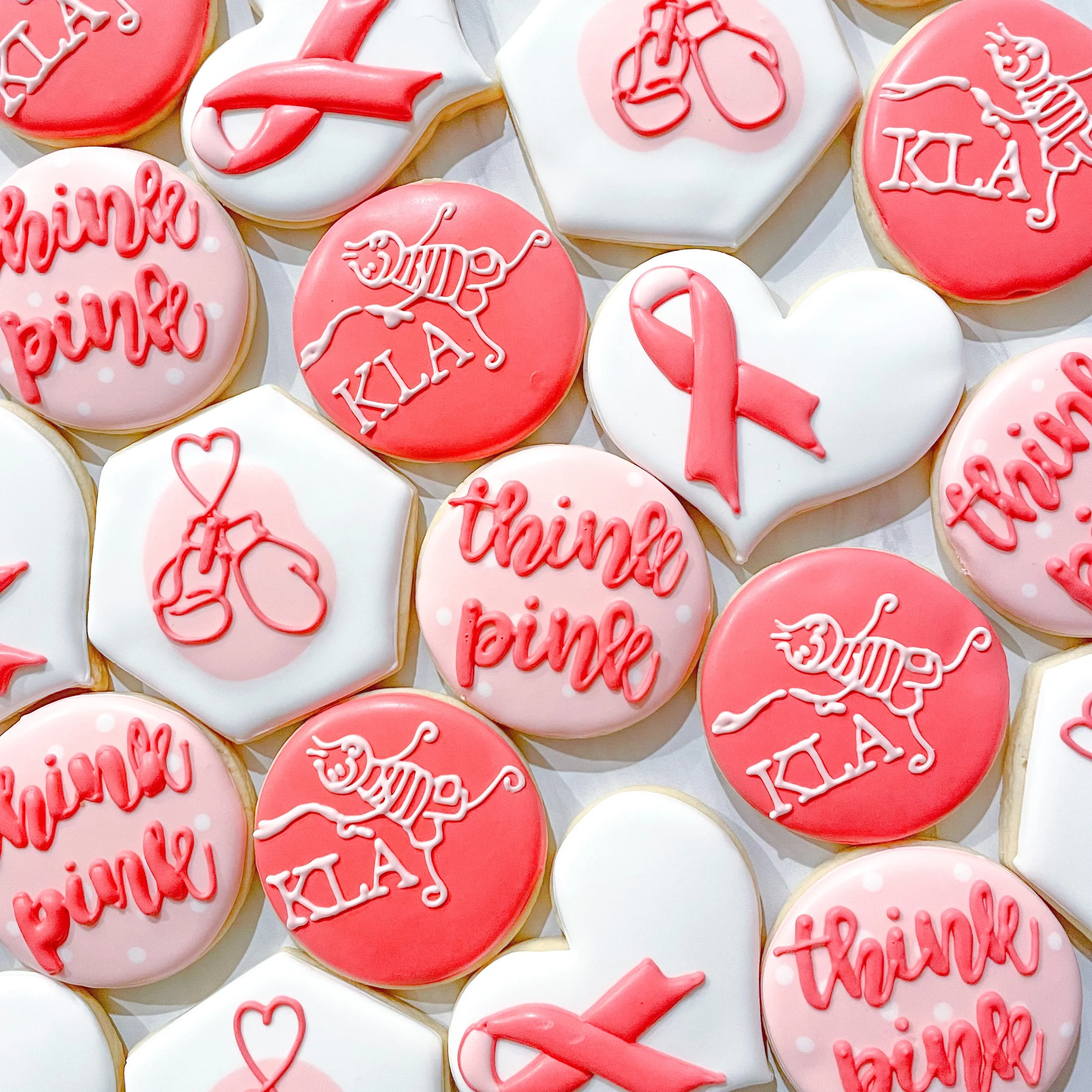 Collection of decorated cookies with pink and white icing, featuring messages such as "Thanks", "love", and symbols like hearts and ribbons, for Breast Cancer Awareness.