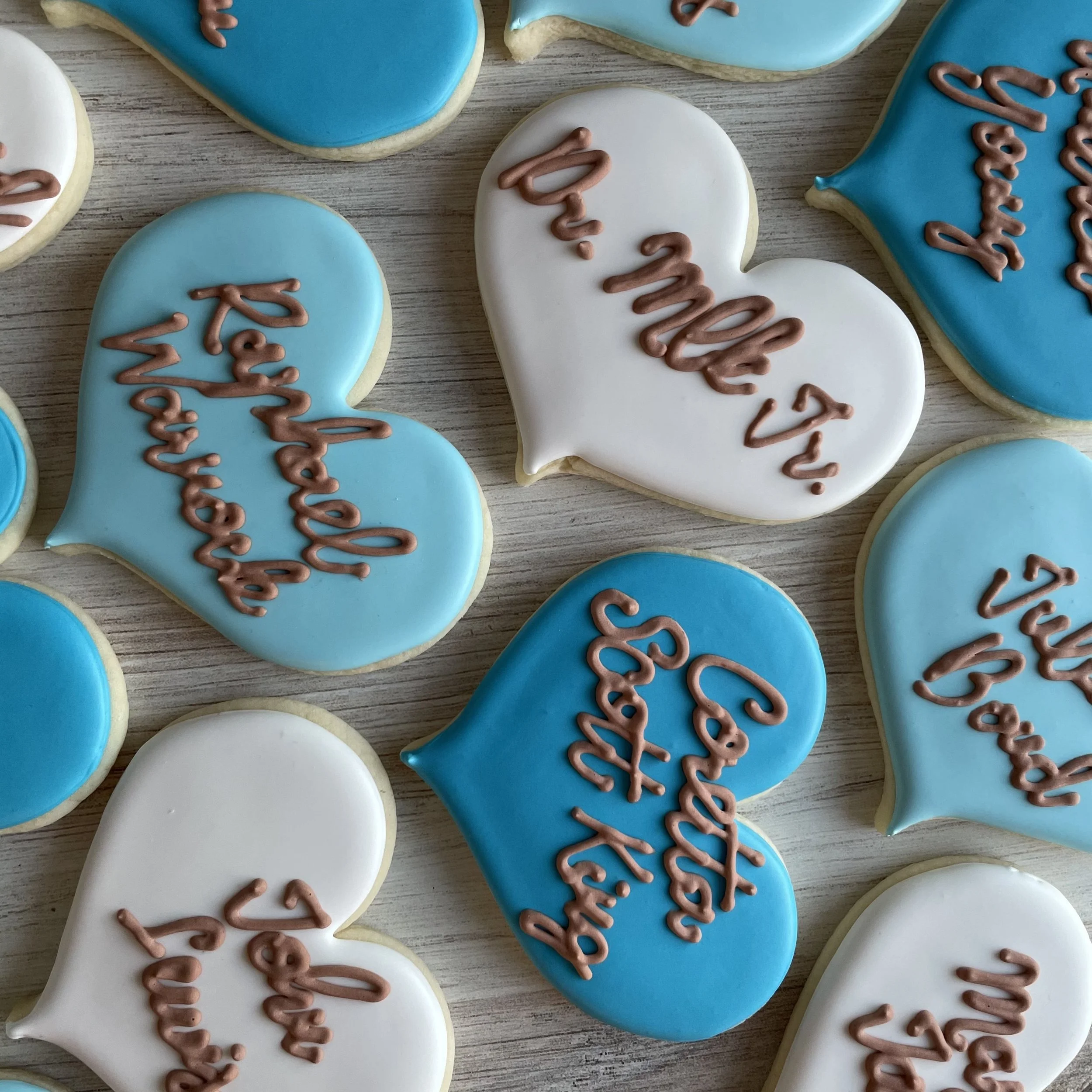 Decorated cookies with blue and white icing that spell out 'SANTA' in cursive pink icing, arranged on a wooden surface.