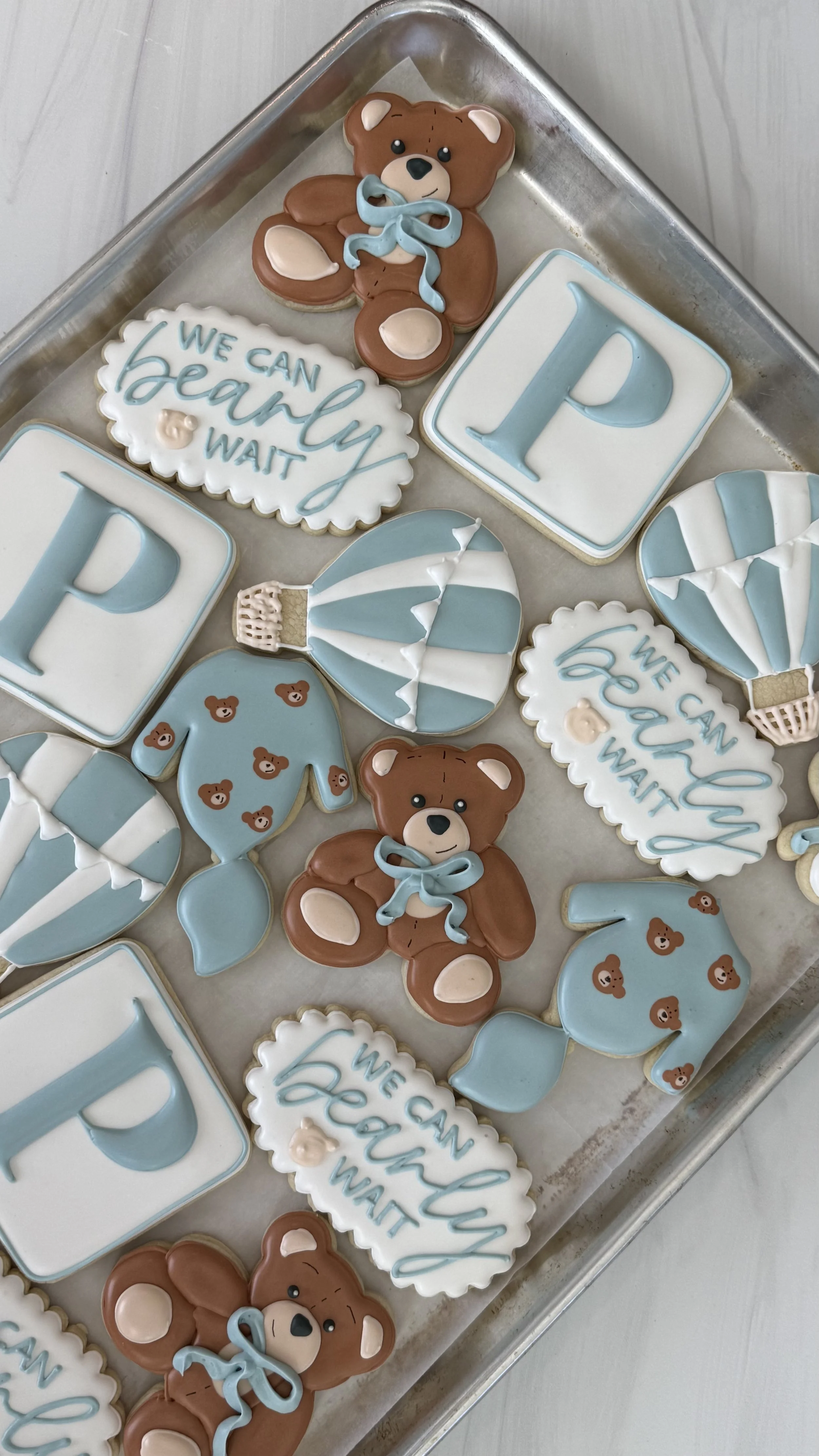 Tray of decorated cookies featuring teddy bears, hot air balloons, and inspirational messages in blue and white colors.