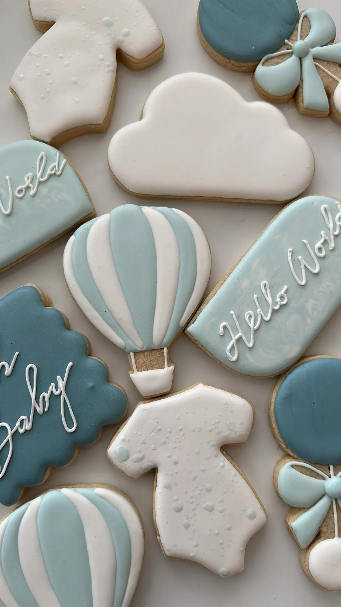 Assorted sugar cookies decorated with light blue, white, and cream-colored icing, with designs including clouds, a hot air balloon, a bow, and the words 'Hello World'.