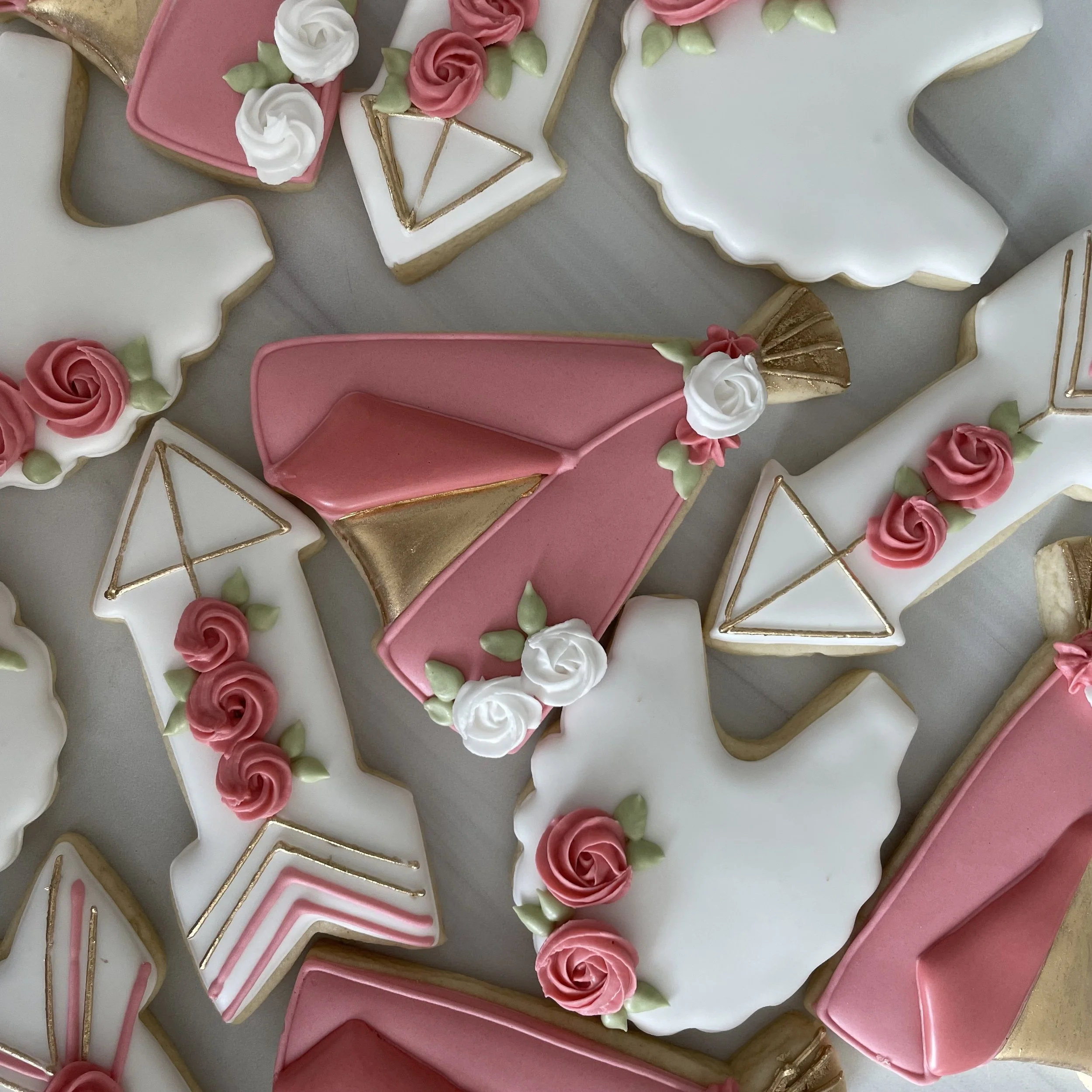 Decorated cookies with pink, white, and gold icing, shaped like hearts, roses, rainbows, and geometric designs.