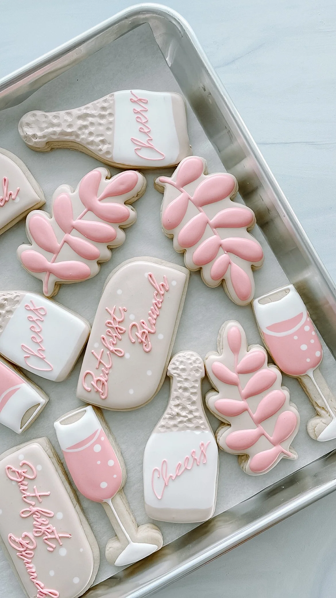 Pink and white decorated cookies on a silver tray, shaped like champagne bottles, glasses, leaves, and with words like 'Cheers' and 'Happy Birthday' in pink icing.