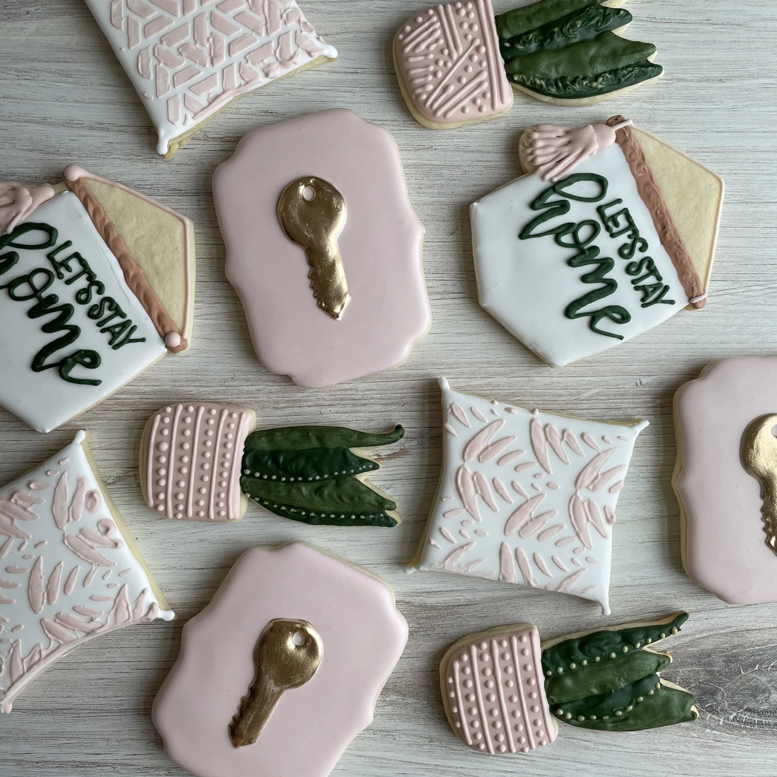 Assorted decorated cookies on a wooden surface, featuring messages like "PESAN WARISAN" and designs such as a green leaf, pink shapes, and gold accents.