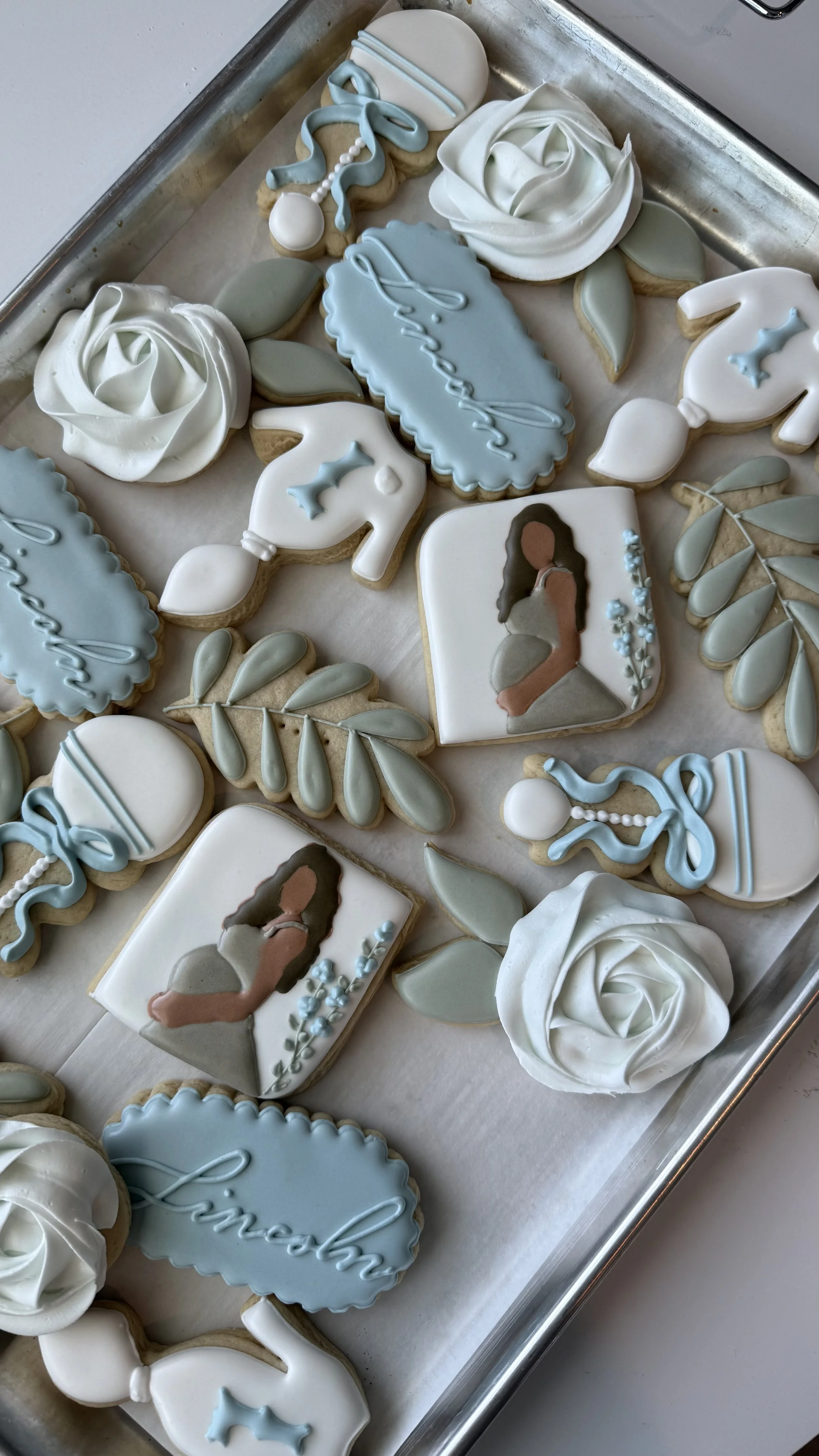 Decorated sugar cookies with baby shower theme, including roses, leaves, a girl silhouette, and phrases like "welcome" and "love" in pastel blue, white, and gray colors.