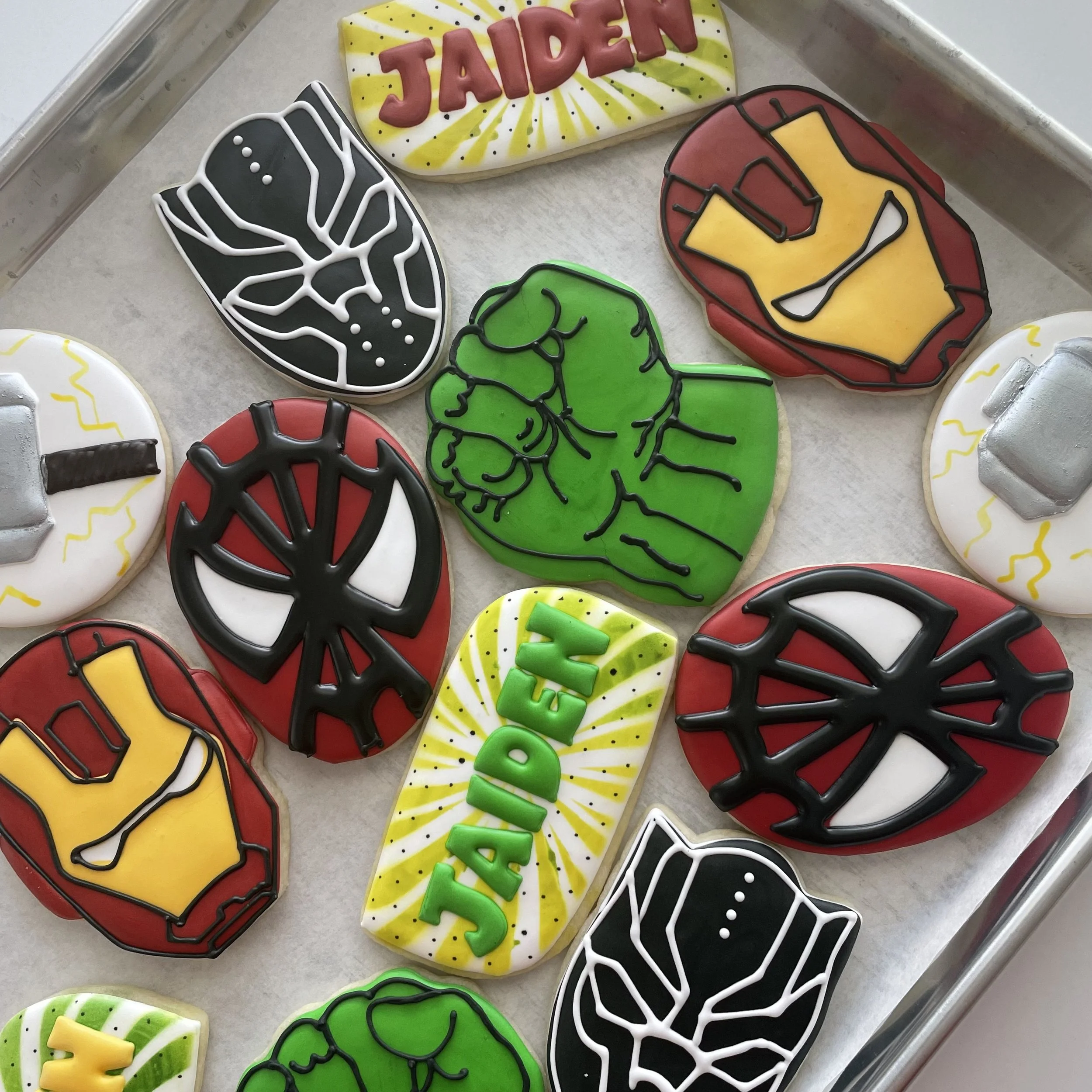 Decorated sugar cookies with a superhero theme, featuring designs such as a black and white spider web, a green superhero mask, a red and yellow shield, a red, yellow, and black helmet, and the words "Jaden" and "Super" in colorful icing.