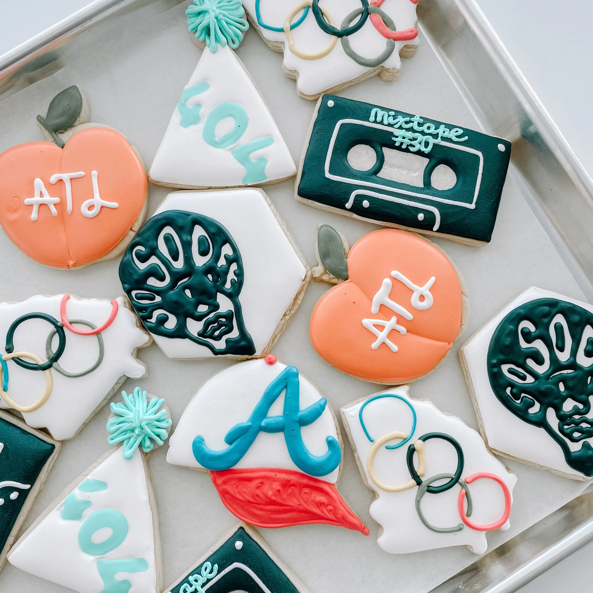 A tray of decorated cookies with summer and mowing themes. Some cookies have phrases like 'I ❤️ ATL' in orange icing, and 'mix tape #30' in teal icing. Others have designs like trees, phones, and abstract lines in various colors such as teal, white, 