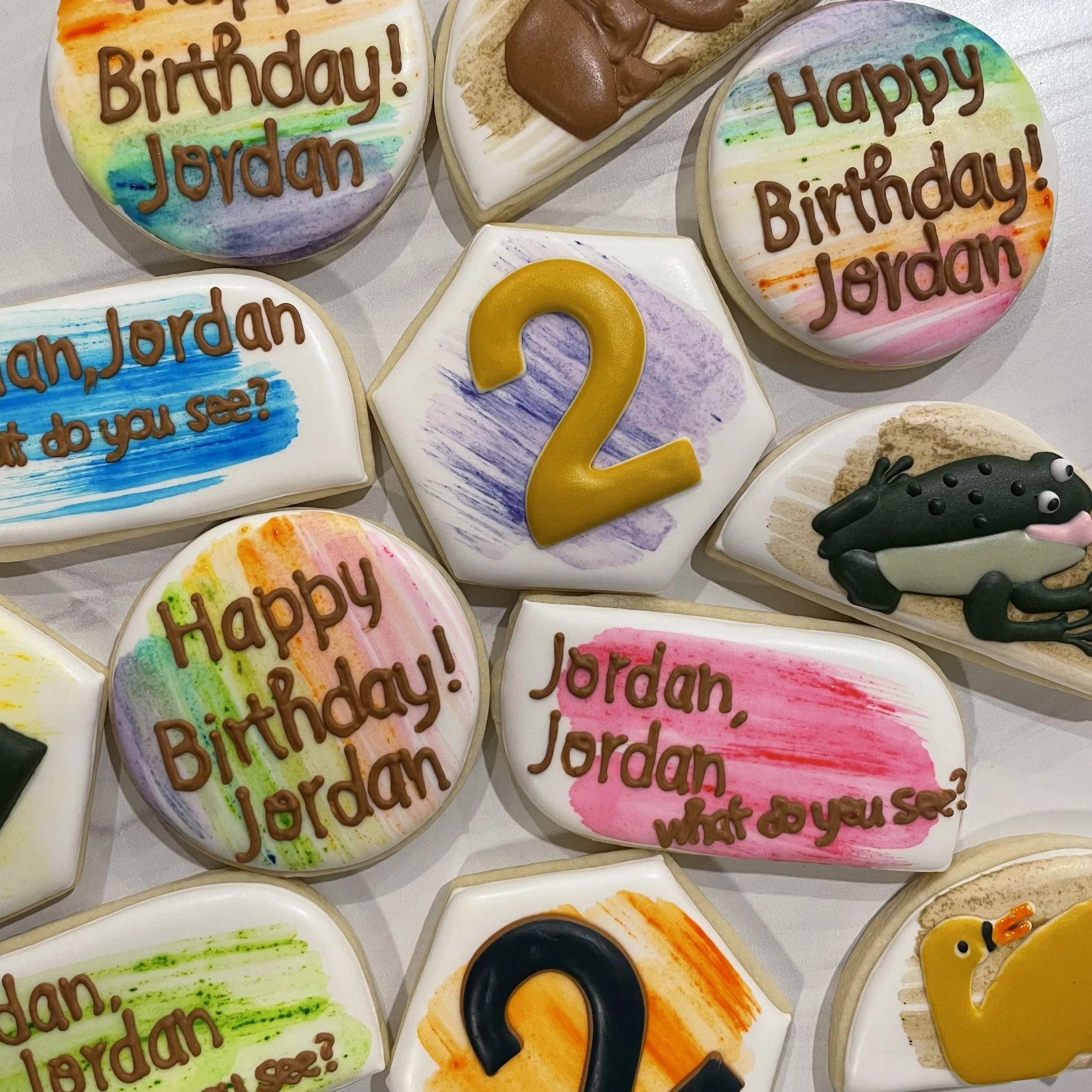 Decorated sugar cookies for a birthday celebration. Some cookies have colorful rainbow backgrounds with the words 'Happy Birthday Jordan' written in chocolate. Others are decorated with various designs, including a big yellow question mark and an ani