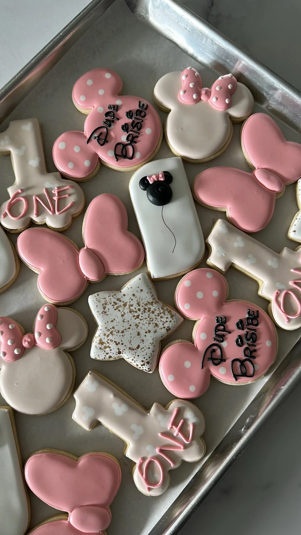 A tray of decorated cookies for a milestone celebration, featuring themed shapes like Minnie Mouse heads, stars, hearts, and bows in pink, white, and black colors with polka dots and writing, including '1' and 'Happy Birthday' messages.