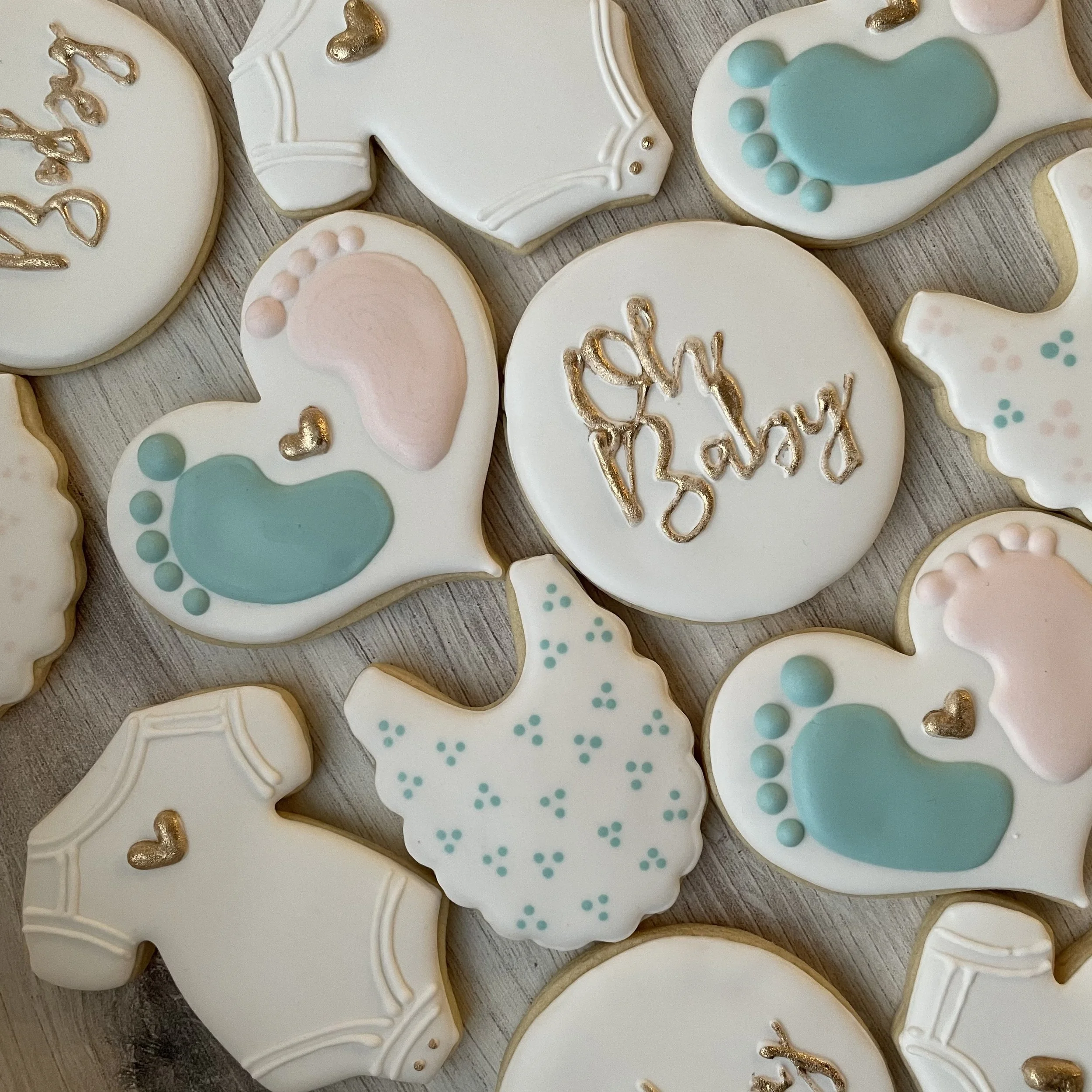Assorted white decorated cookies with pastel and gold accents, some with baby-themed designs, on a wooden surface.