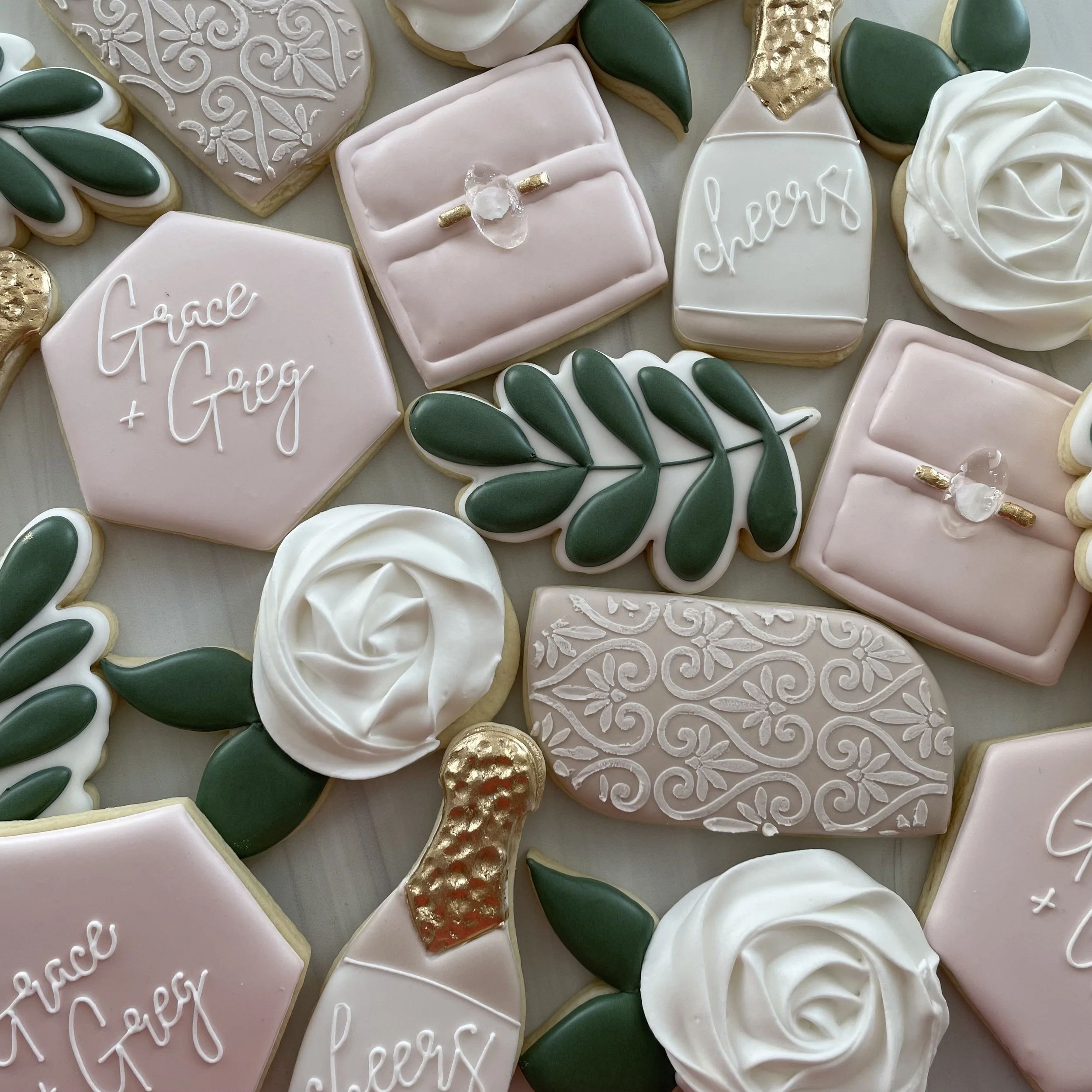 Decorated sugar cookies with a pale pink, white, and gold color scheme. Some cookies have the words "peace" and "happy" written in cursive icing, and others are decorated with green leaves and intricate white floral patterns.