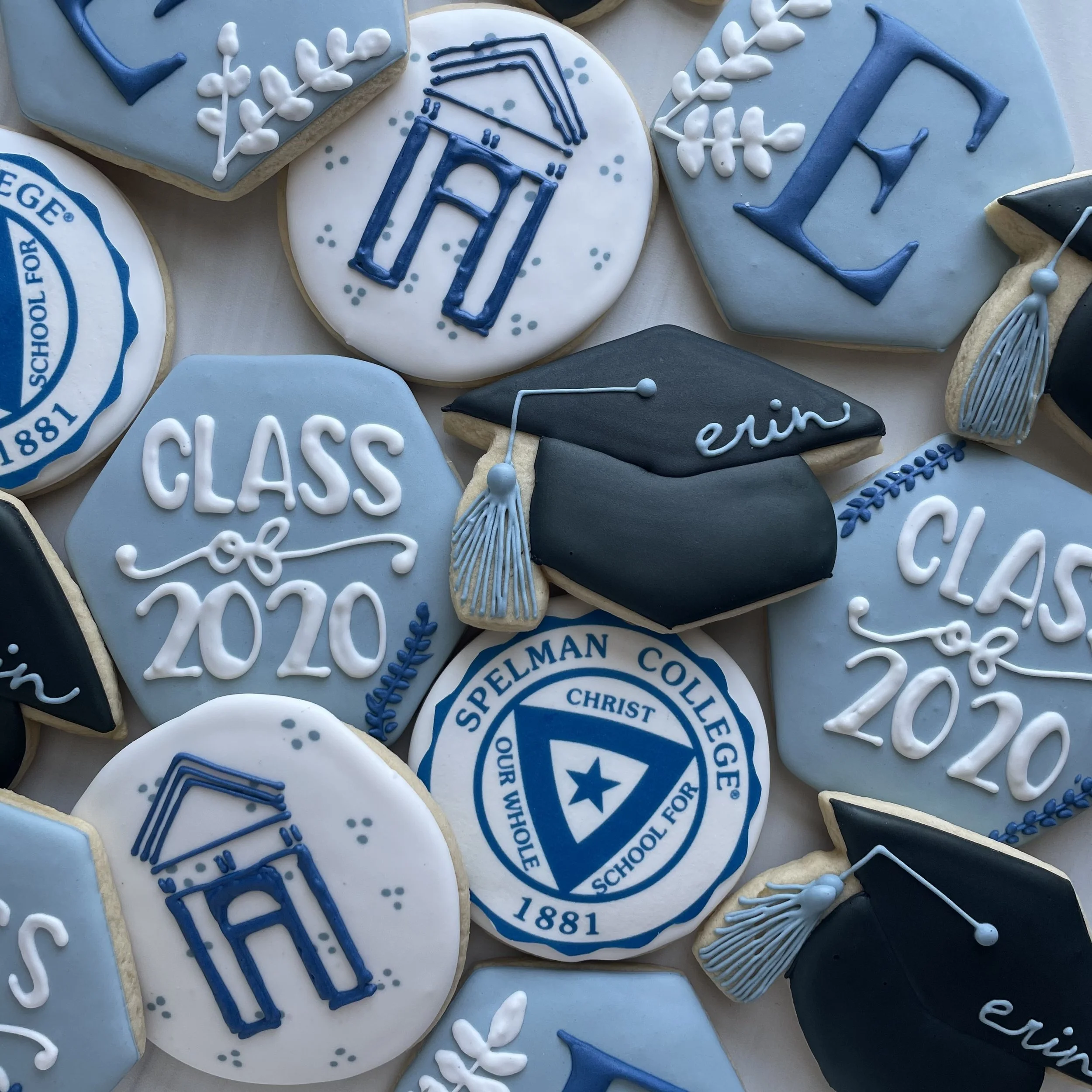 A collection of decorated cookies themed for a school or academic graduation, featuring blue, white, and black icing with academic symbols, school logos, and celebratory words.