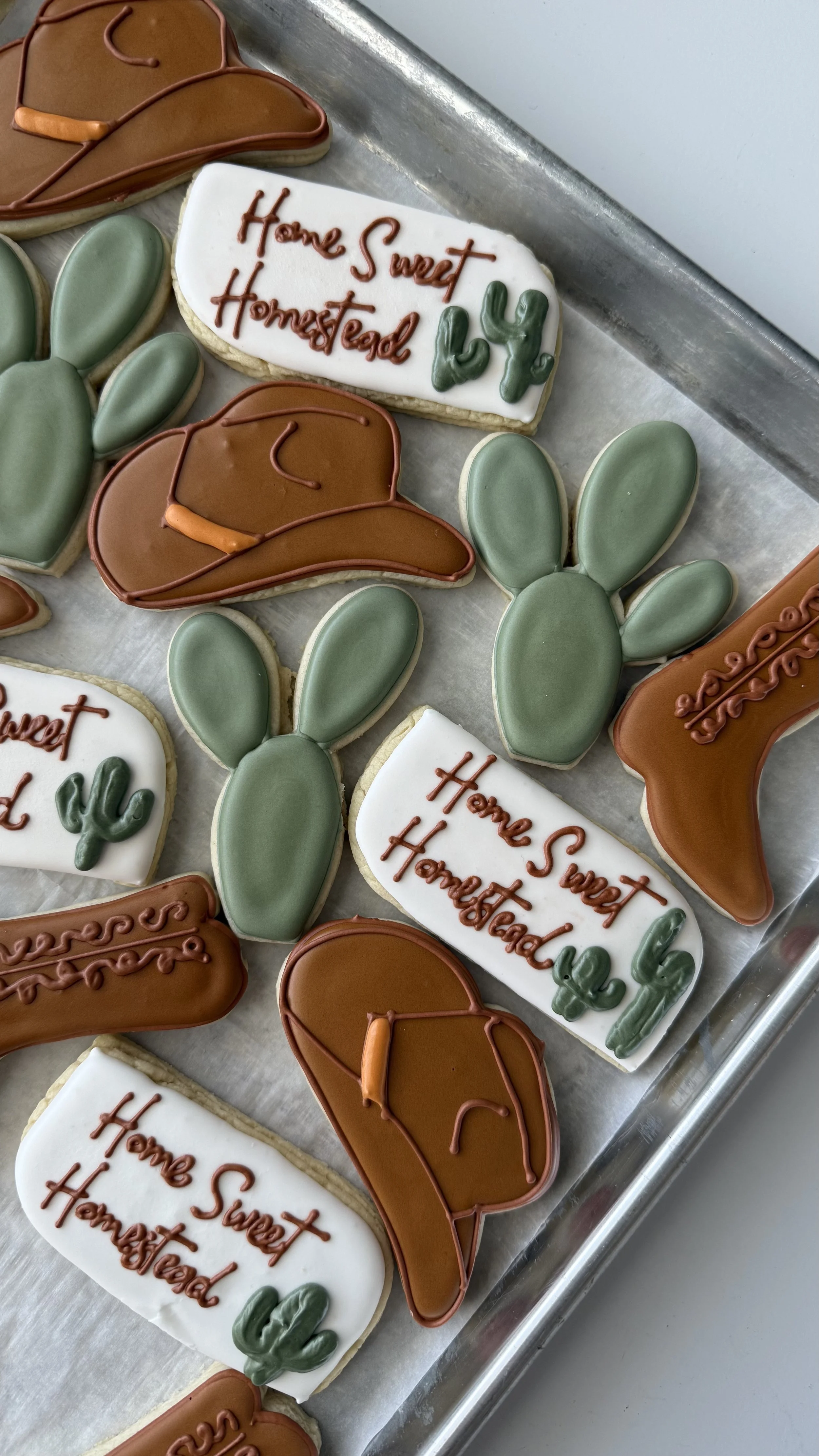Decorative cookies shaped like cacti, cowboy hats, and boots, with a white cookie that reads "Home Sweet Homestead" and green cactus decorations.